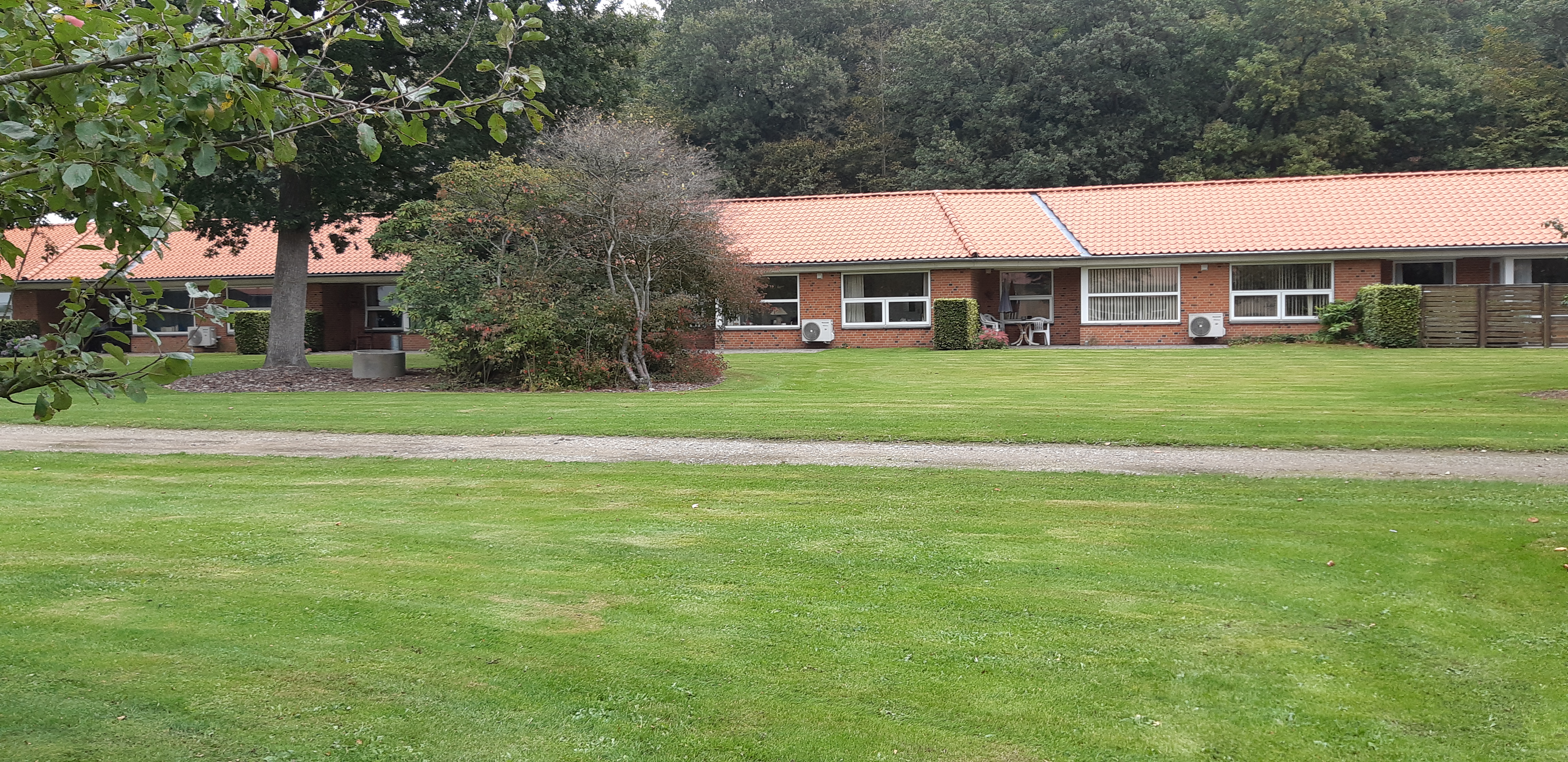 Solgården set ude fra, bygningen er en lang og der er røde mursten og hvide vinduer. Der er en stor flot grøn græsplæne og to store træer.