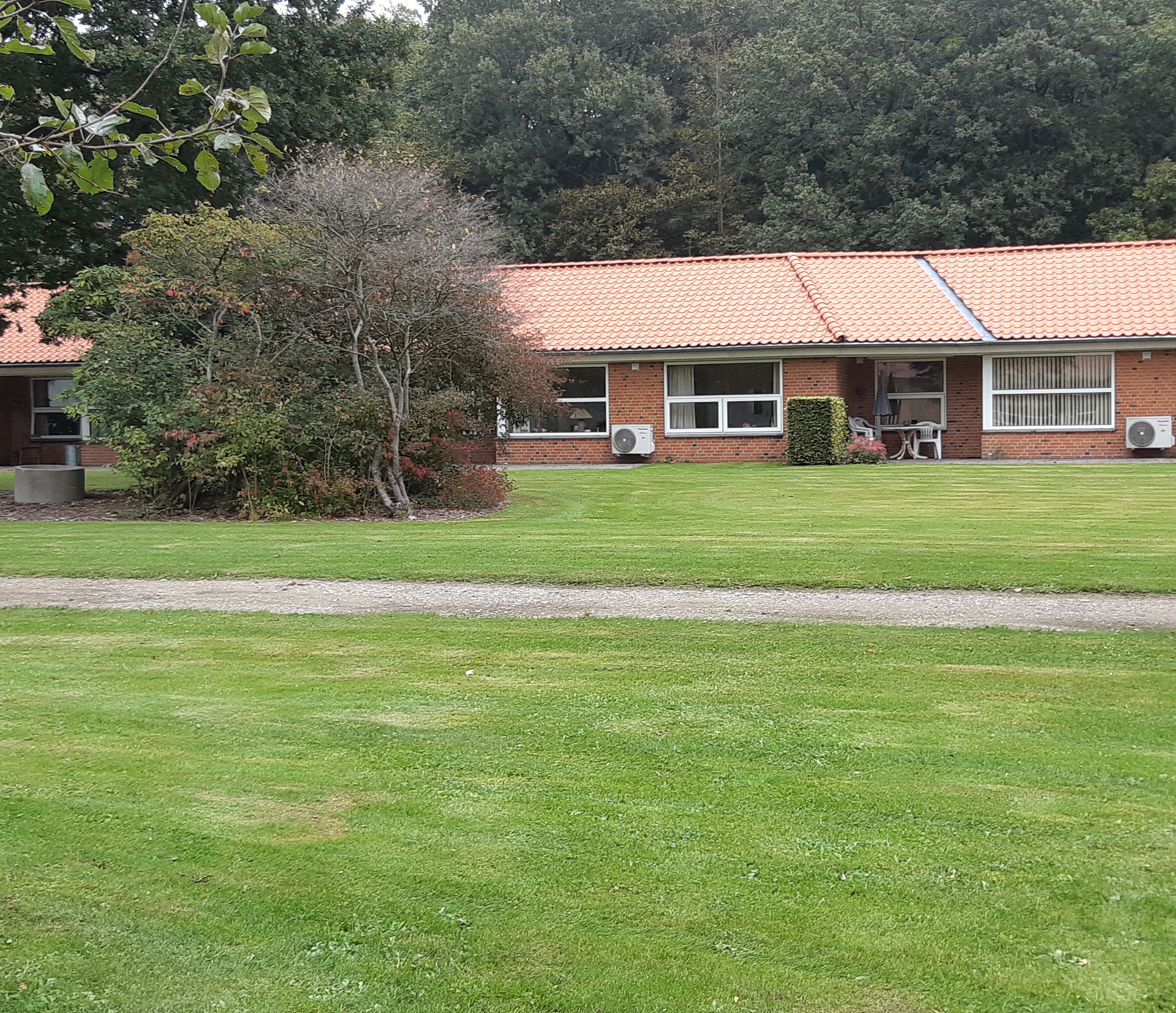 Solgården set ude fra, bygningen er en lang og der er røde mursten og hvide vinduer. Der er en stor flot grøn græsplæne og to store træer.