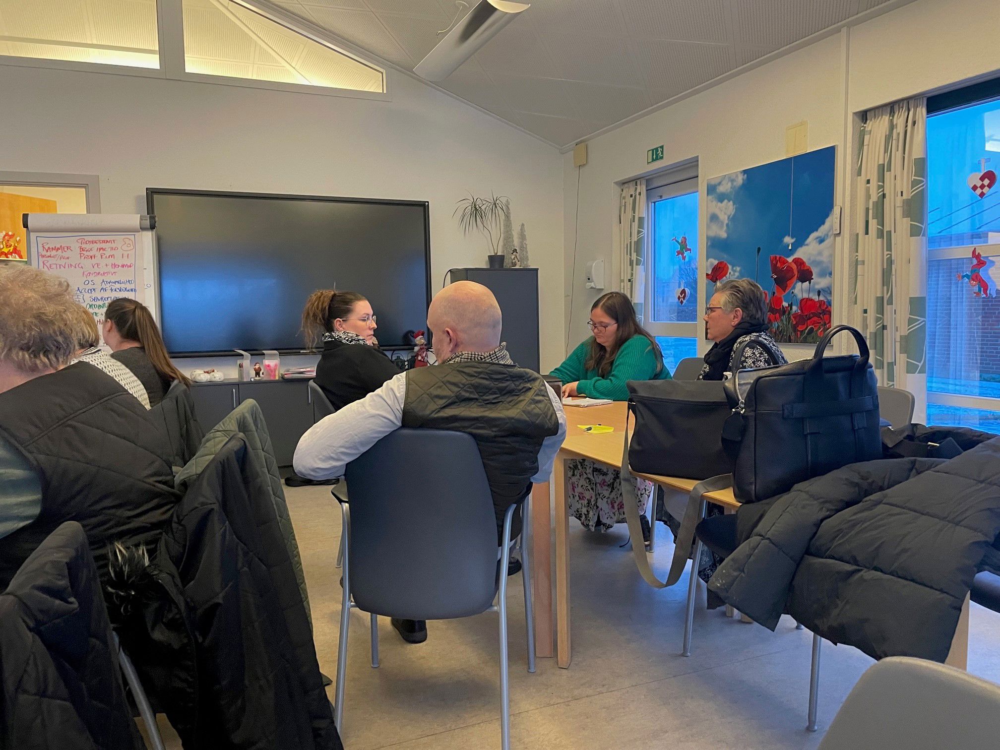 På billedet ses et bord hvor der sidder 4 personer de snakker sammen og en skriver notater. Bagerst i rummet er der en flipover og en stor skærm. I vinduet hænger julepynt.
