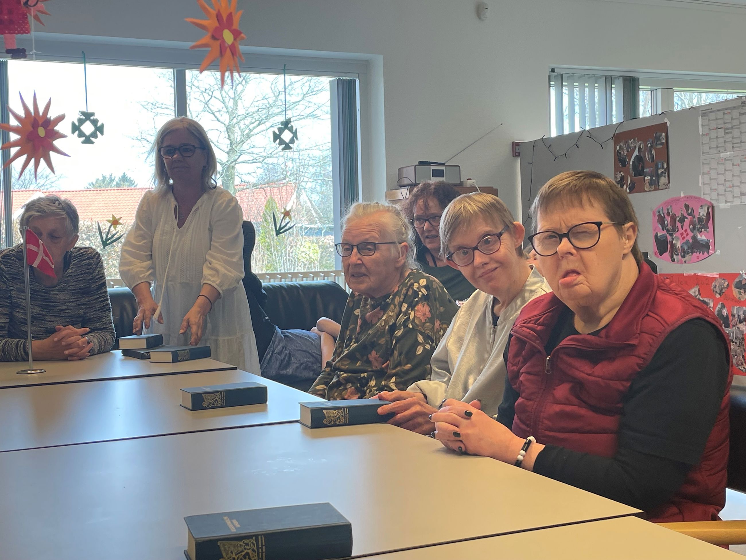 Nogle borgere sidder rundt om et bord foran dem ligger en blå sangbog fra folkehøjskolen, nederst på bordet står et flag foran en borger, ned fra loftet hænger der orange blomster
