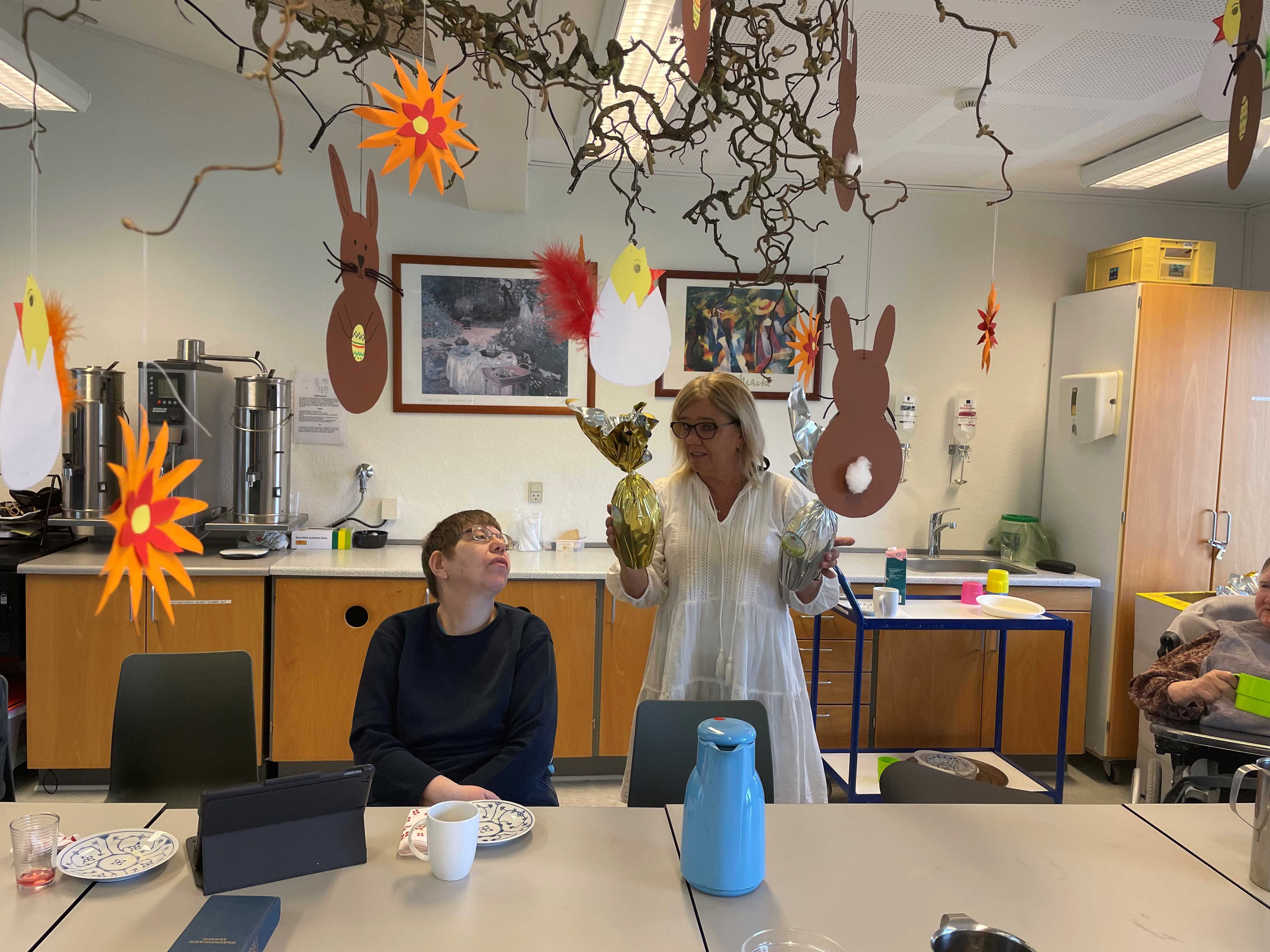 To borgere sidder ved et bord mens en ansat står med to påskeæg og er ved at fortælle noget, i loftet hænger udklippet påskepynt på en troldhasselgren