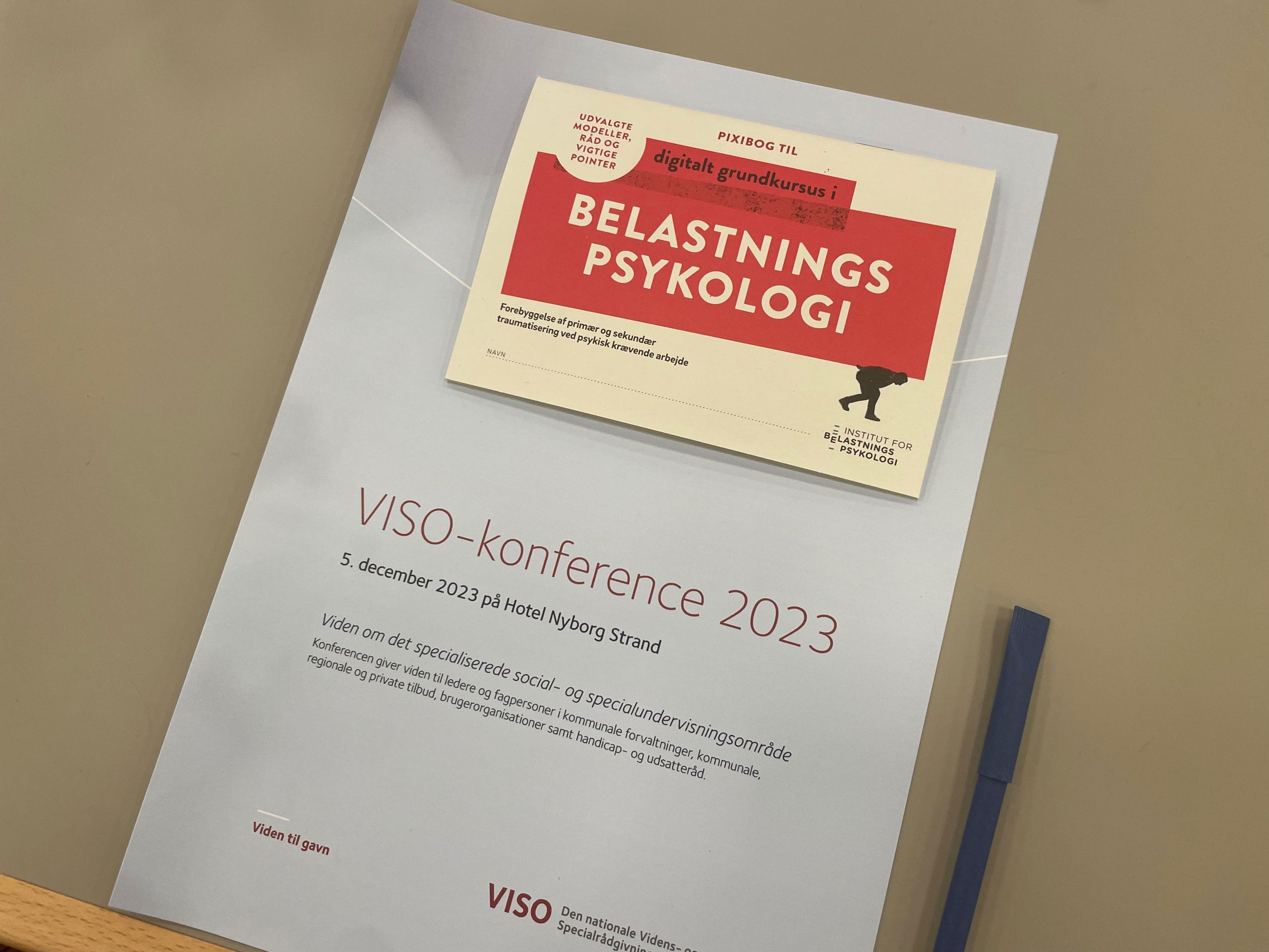 En grå mappe med indhold om hvad der skal ske på VISO konference der bliver afholdt den 5. december 2023 på Hotel Nyborg Strand, ligger på et bord sammen med en kuglepen.
