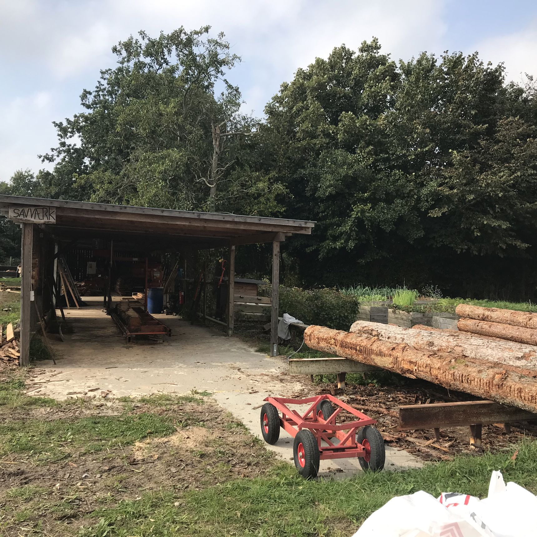 Træværksted hvor der til højre i billedet ligger seks trærafter og bagerst i billedet ligger træværkstedet, det er bygget i mørkt træ.