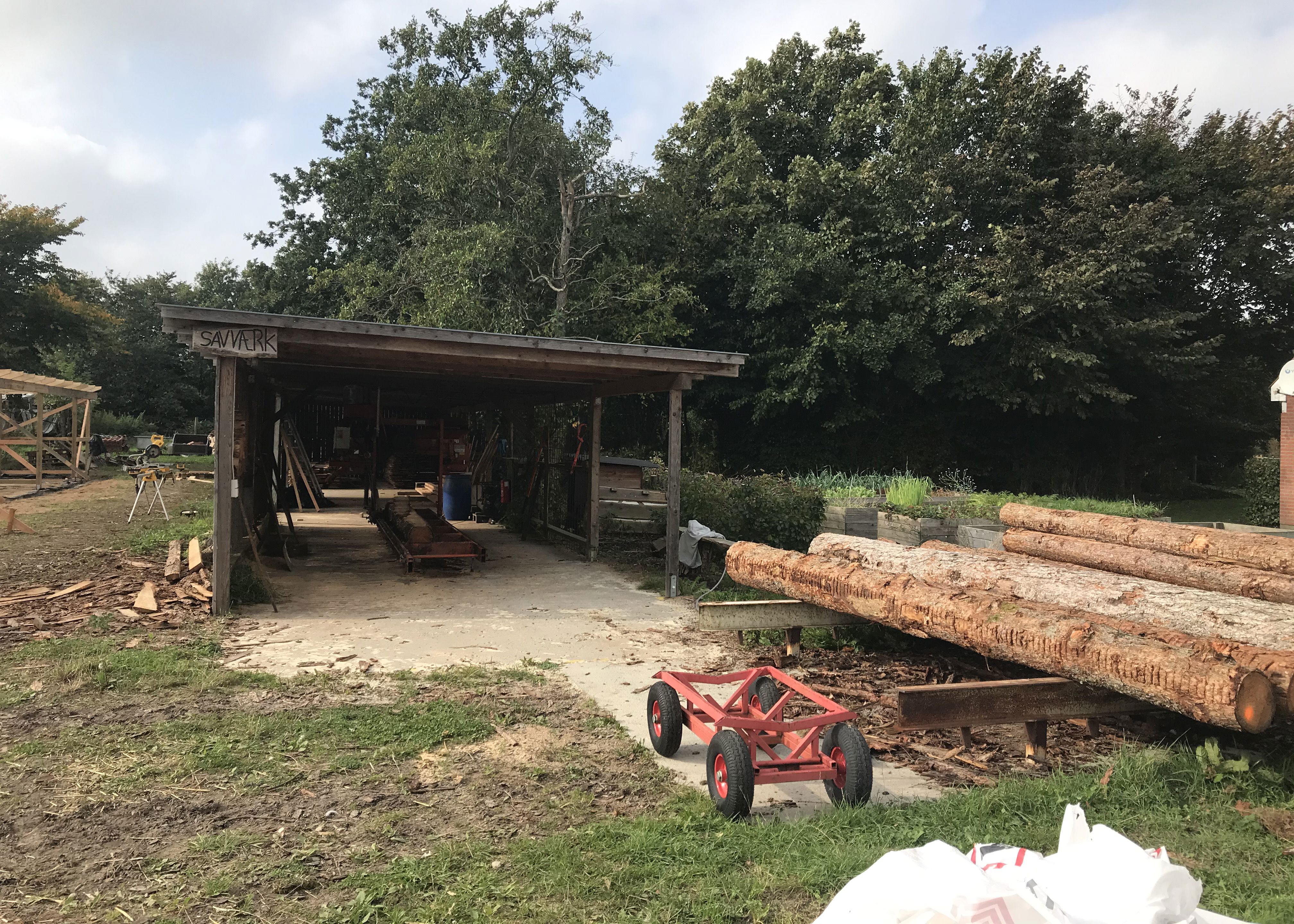 Træværksted hvor der til højre i billedet ligger seks trærafter og bagerst i billedet ligger træværkstedet, det er bygget i mørkt træ.