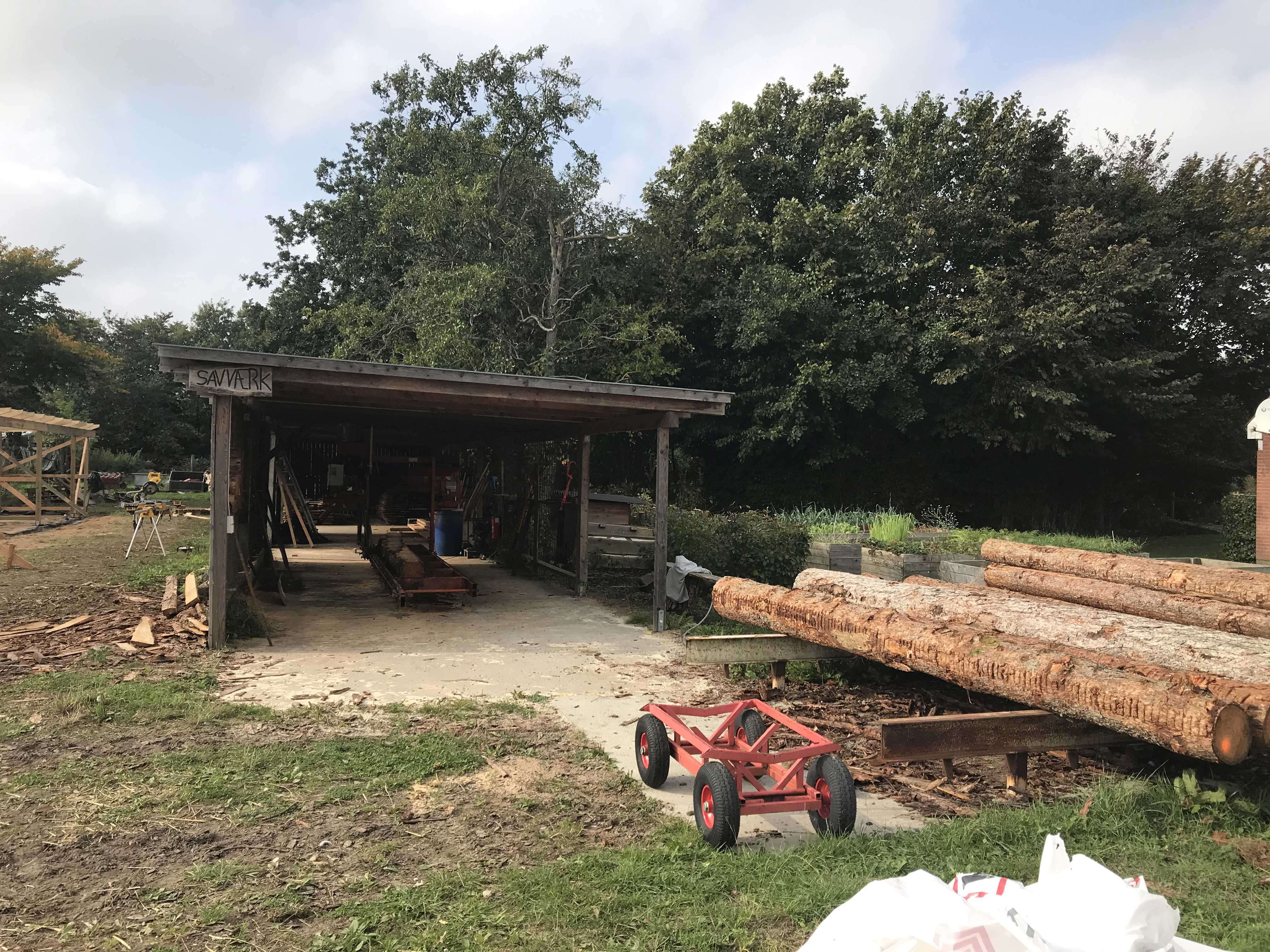 Træværksted hvor der til højre i billedet ligger seks trærafter og bagerst i billedet ligger træværkstedet, det er bygget i mørkt træ.