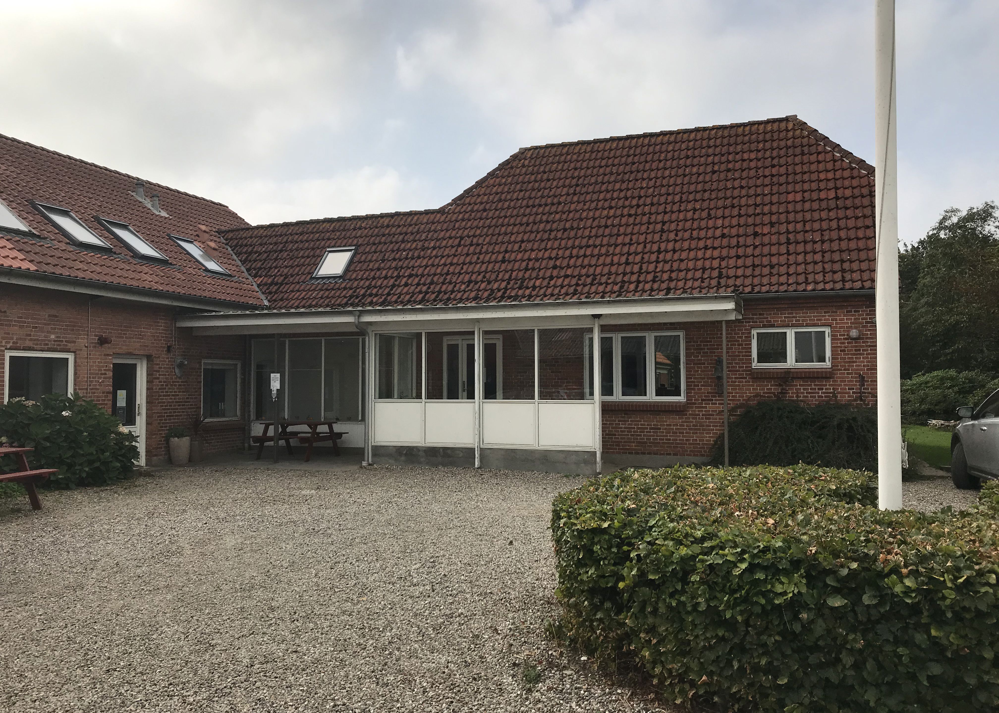 hovedbygning i røde sten og rød tegltag, indgang med glasparti foran, en gårdsplads med grus og i midten står der en flagstang og rundt om den en lav grøn hæk.