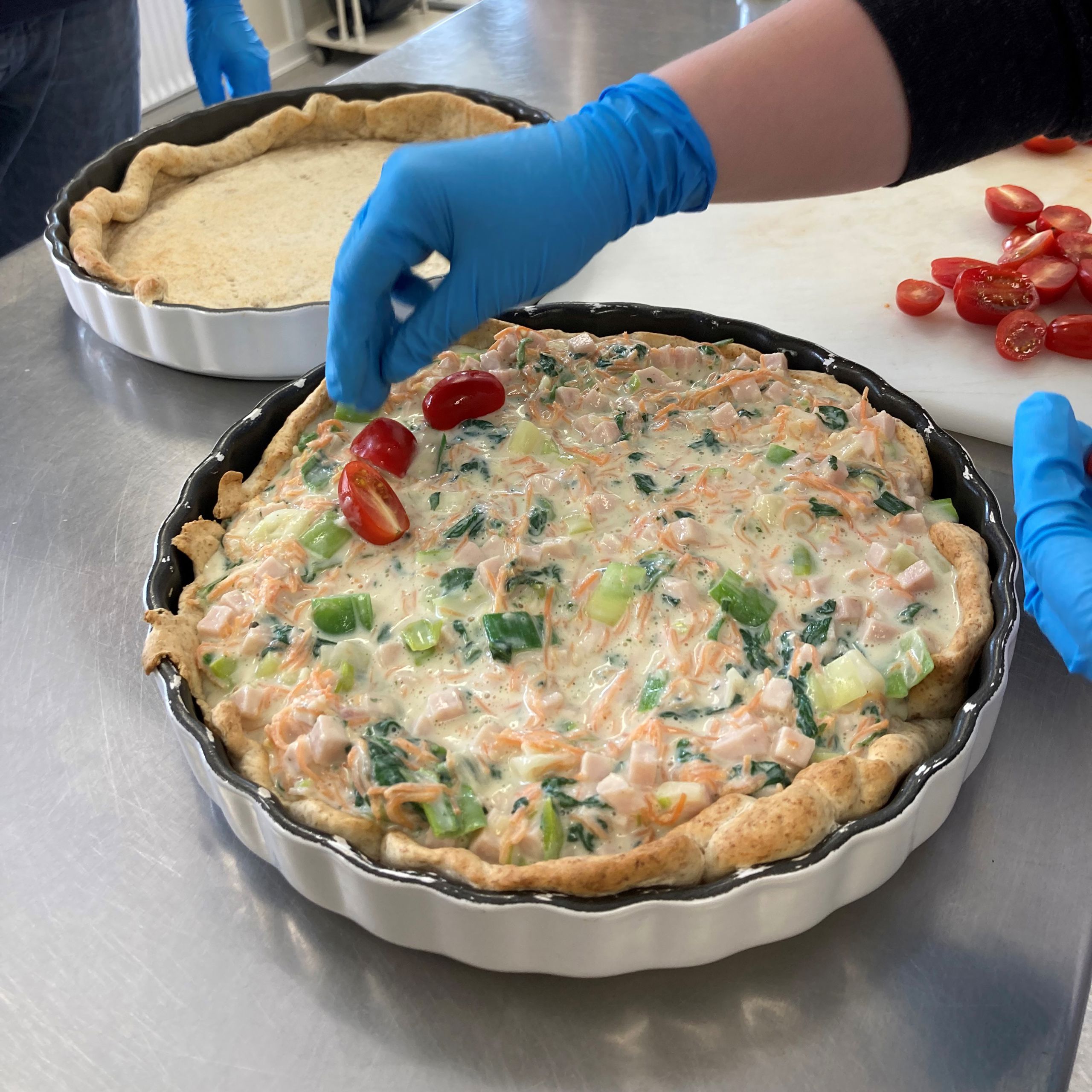 Tomaterne ligges ovenpå tærten ved siden af ses en tæret uden fyld, endnu bagved står andre borgere og kigger på.