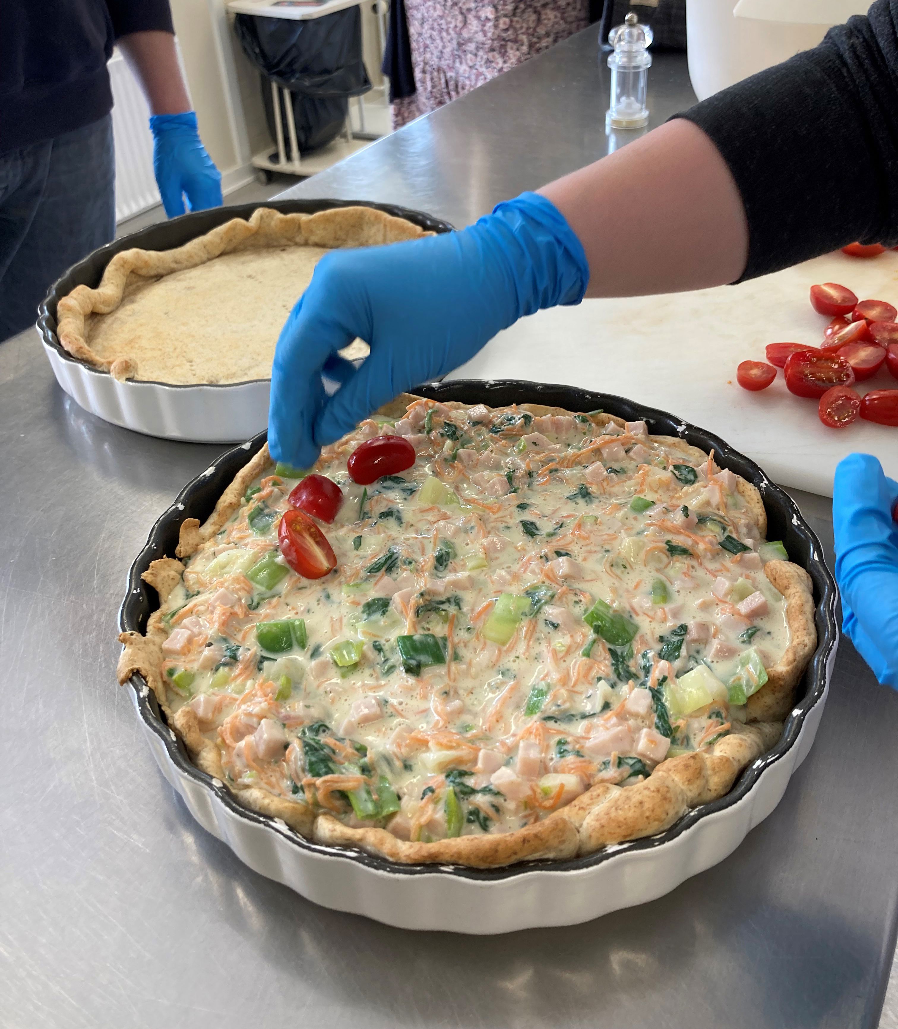 Tomaterne ligges ovenpå tærten ved siden af ses en tæret uden fyld, endnu bagved står andre borgere og kigger på.