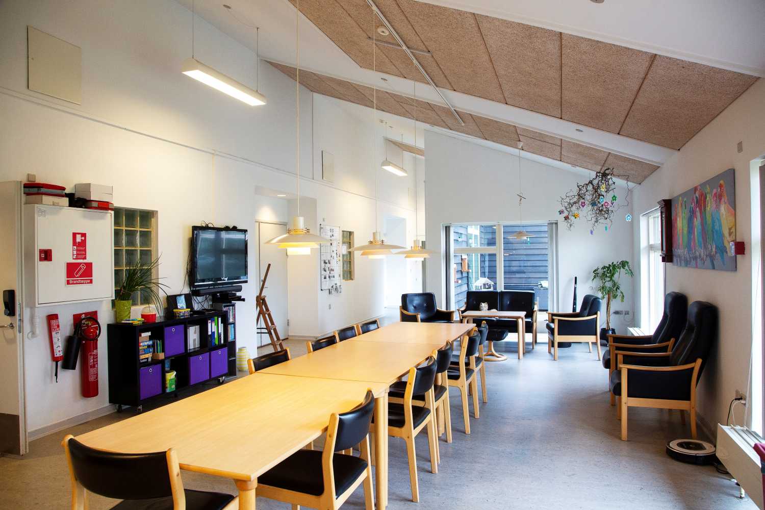 Fællesrum og midt i rummet står der et stort langt bord med stole omkring. På væggen til højre hænger der et tv og der er billeder på væggene.