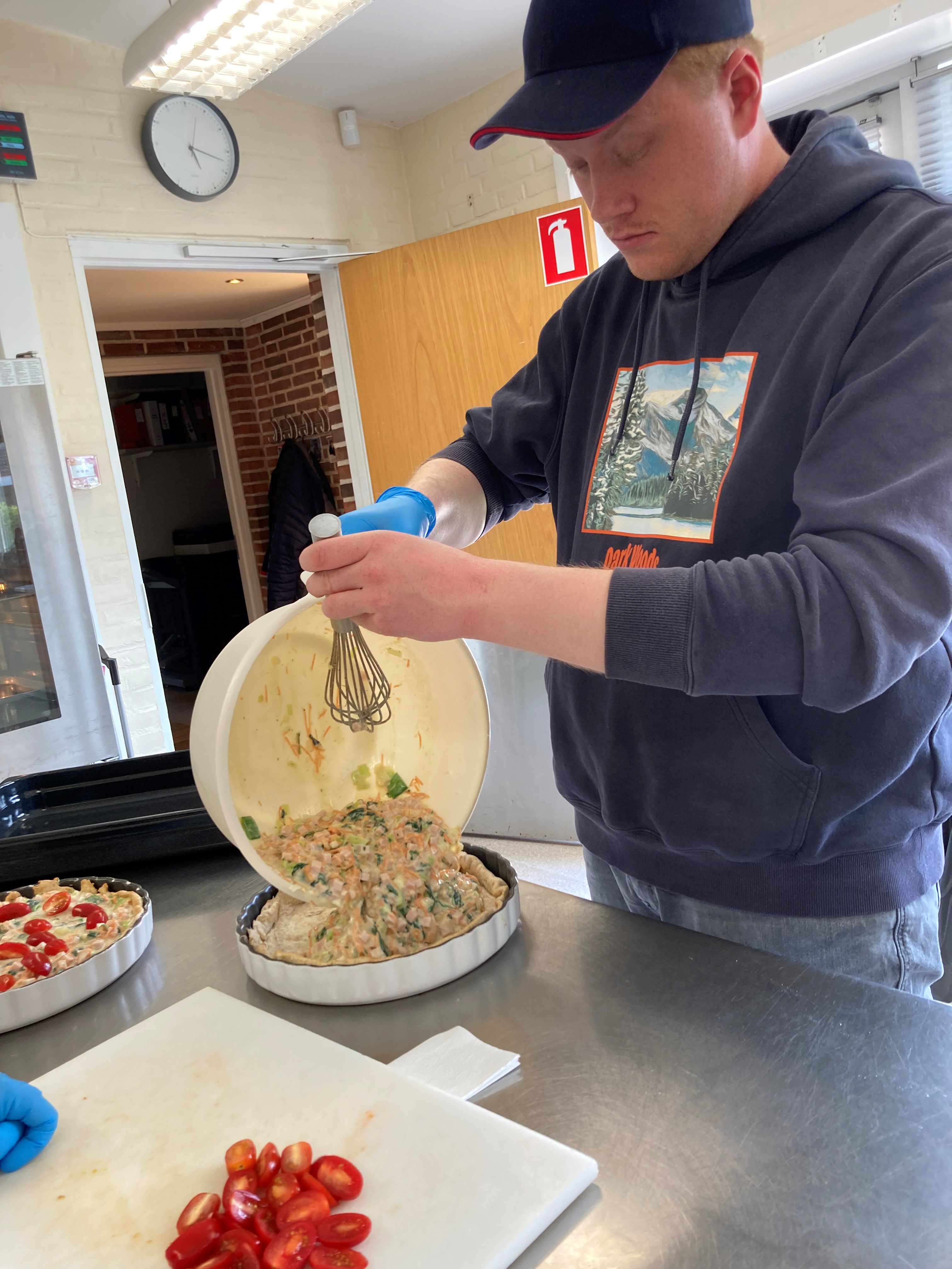 En borger står ved et tærtefad og hælder fyld af kød æg og porre ned i, ved siden af ham på skærebrættet ligger de halve tomater lige til at ligge ovenpå.