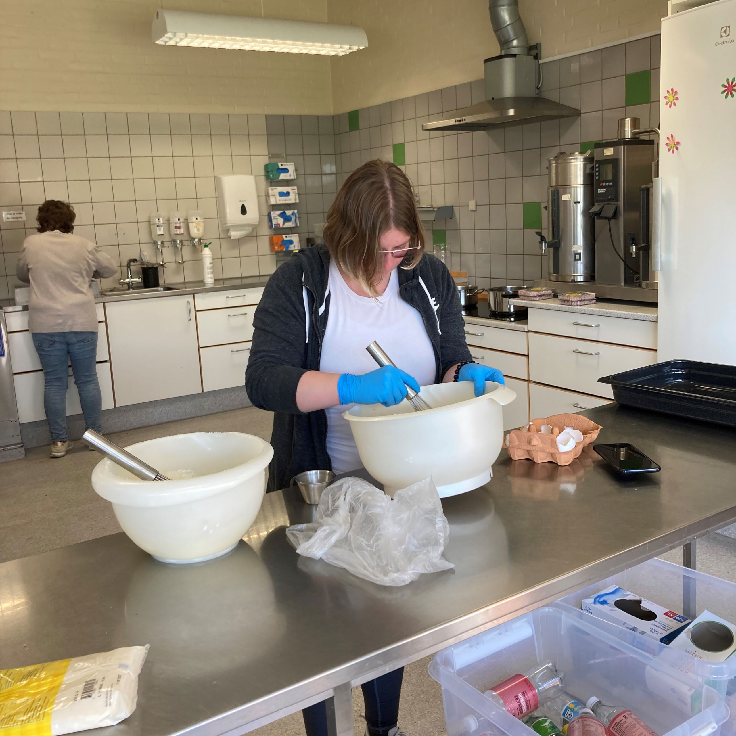 En borger står ved et stort bord og pisker æg sammen i stor skål, bag hende ser man en anden borger står ved vasken, på væggen på den anden side hænger der handsker i alle størrelser.