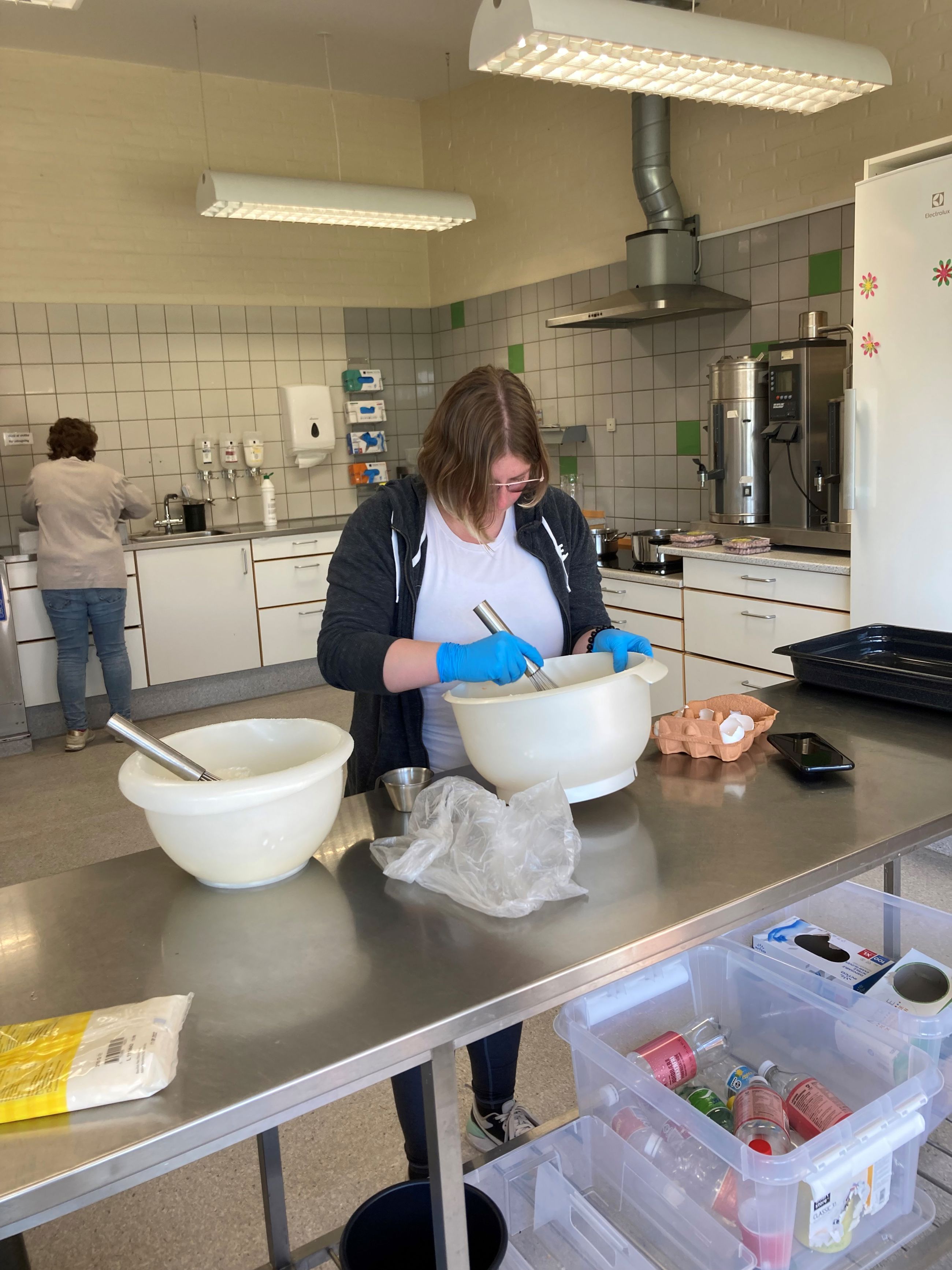 En borger står ved et stort bord og pisker æg sammen i stor skål, bag hende ser man en anden borger står ved vasken, på væggen på den anden side hænger der handsker i alle størrelser.