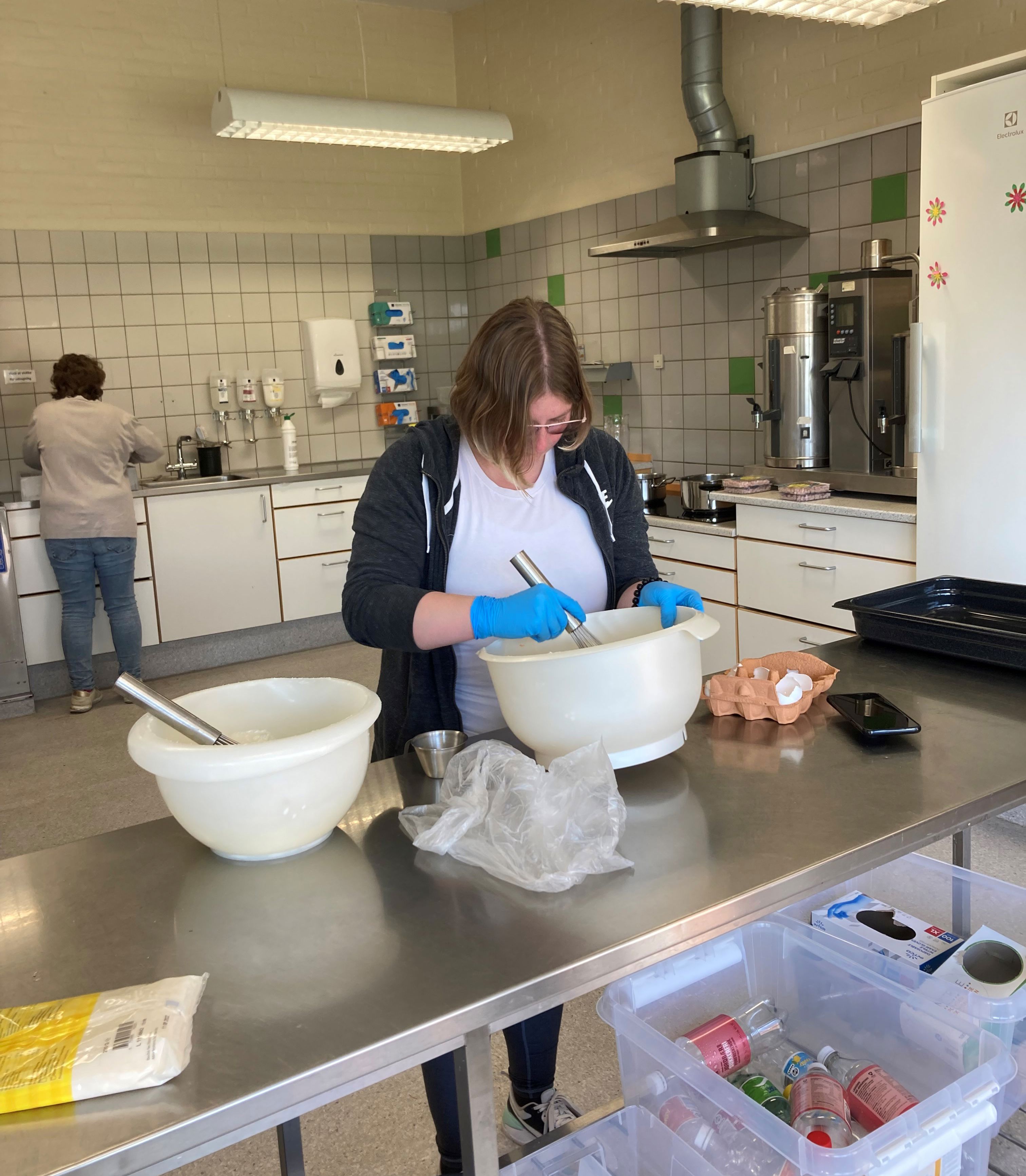 En borger står ved et stort bord og pisker æg sammen i stor skål, bag hende ser man en anden borger står ved vasken, på væggen på den anden side hænger der handsker i alle størrelser.