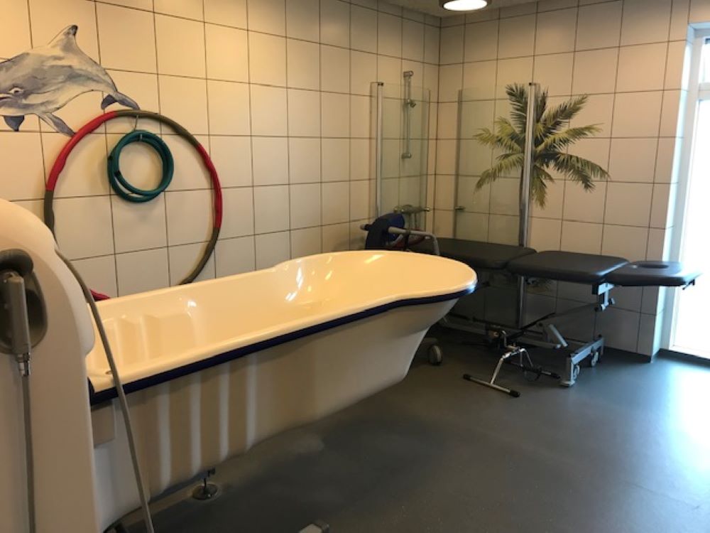 Dette er et badeværelse, hvor der i forgrunden står et hvidt badekar. Gulvet er sort linoleum og der er hvide klinker på væggene.