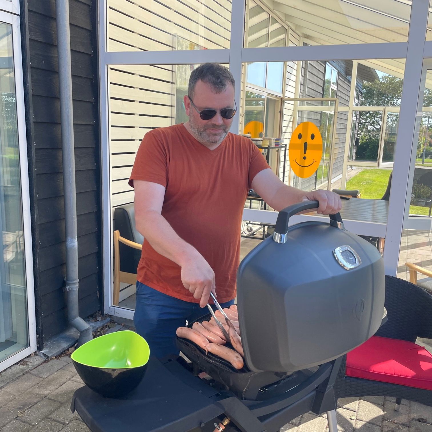 Man ser en gasgrill med fyldt op med pølser der er ved at blive grillet, der står en mand og styrer det,