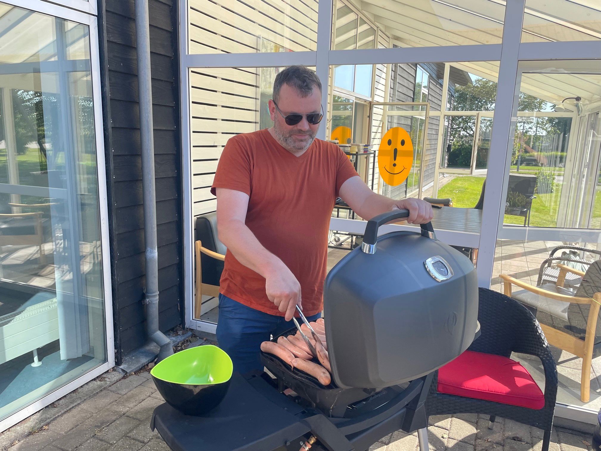 Man ser en gasgrill med fyldt op med pølser der er ved at blive grillet, der står en mand og styrer det,