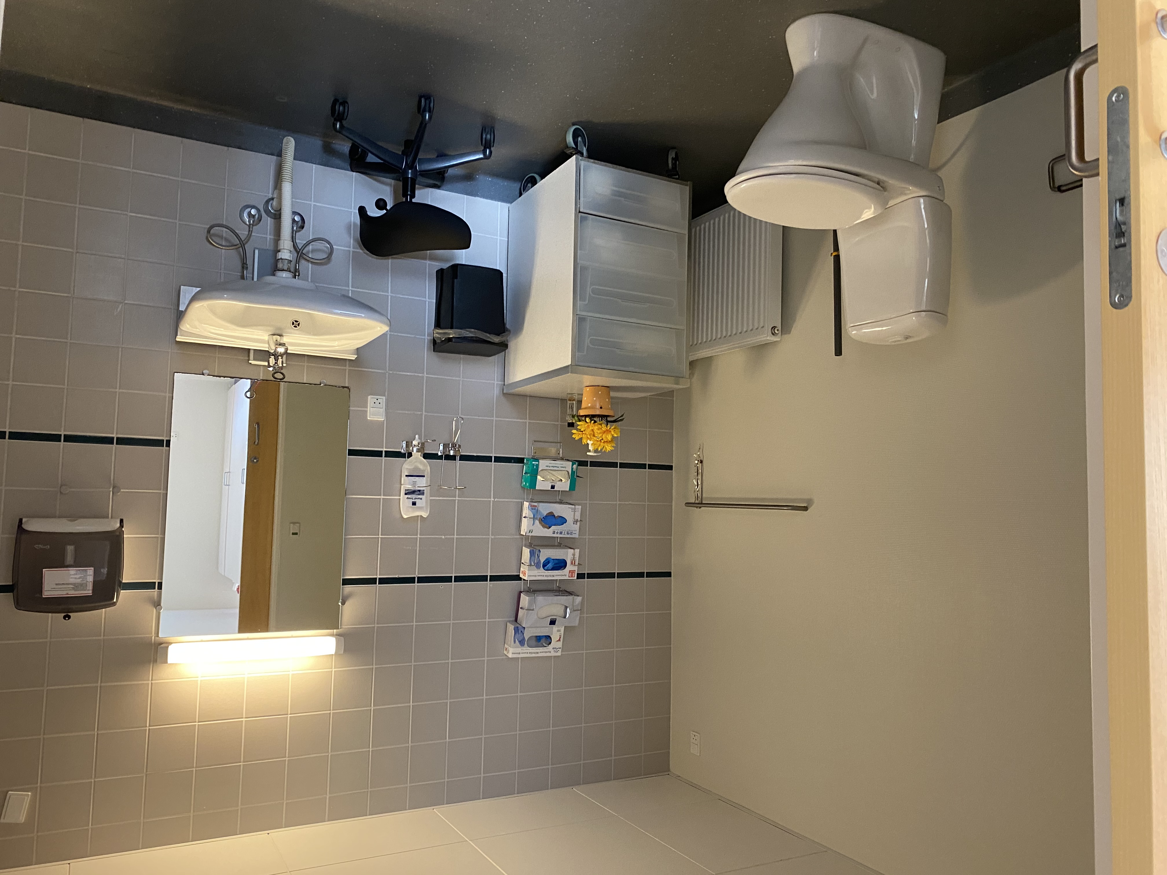 Toilet i borgers lejlighed. Der er et toilet, håndvask og et spejl. I venstre hjørne er der en komode med fire skuffer. Gulvet er mørkegråt. 