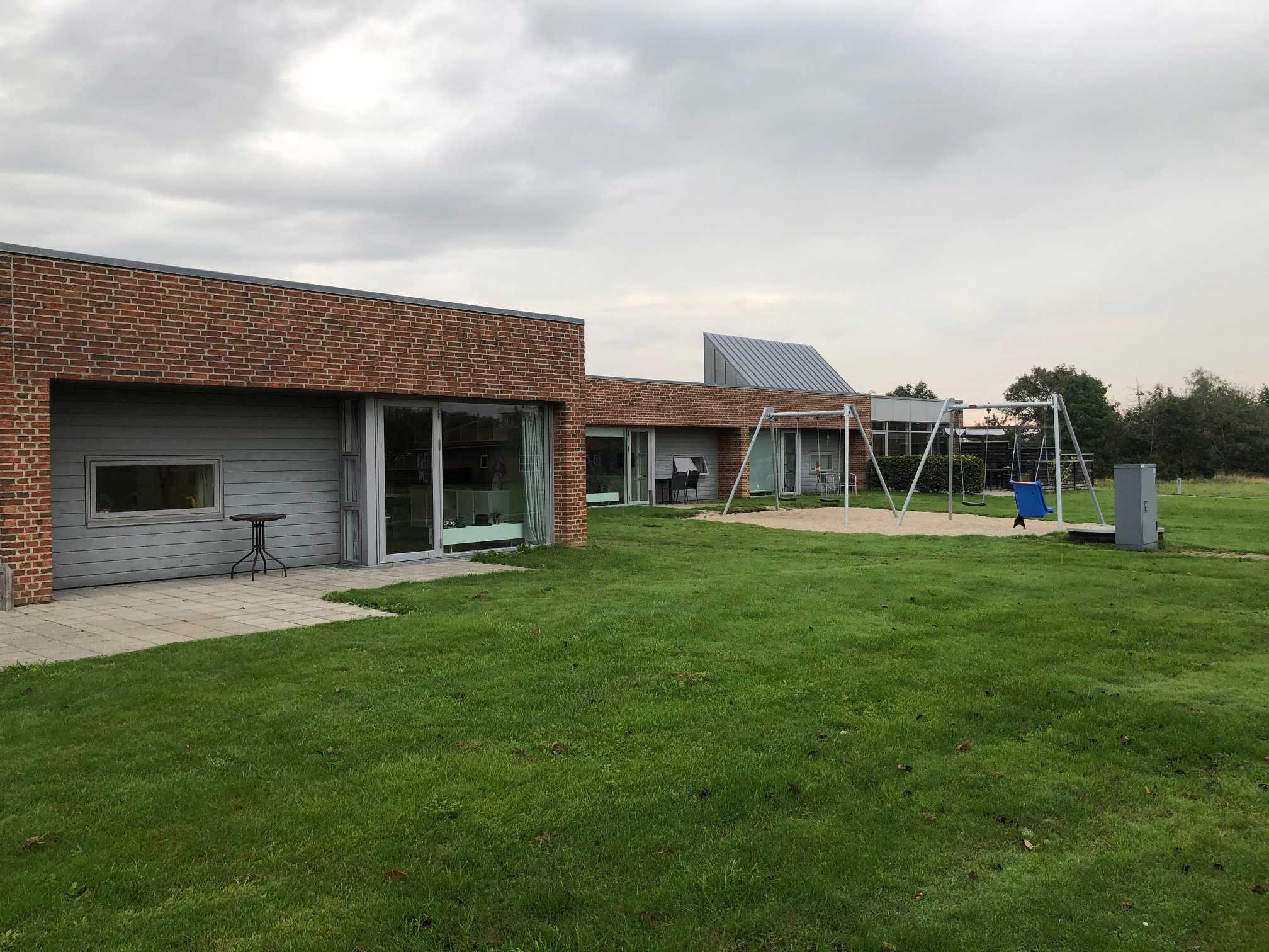 Æblevej udenfor, bygningerne er af røde mursten, og træ der er malet gråt. Foran bygninger er der en grøn græsplæne, og til højre i billedet er der to gyngestativer med hver to gynger på. Gyngestativerne er placeret ovenpå et område med sand.