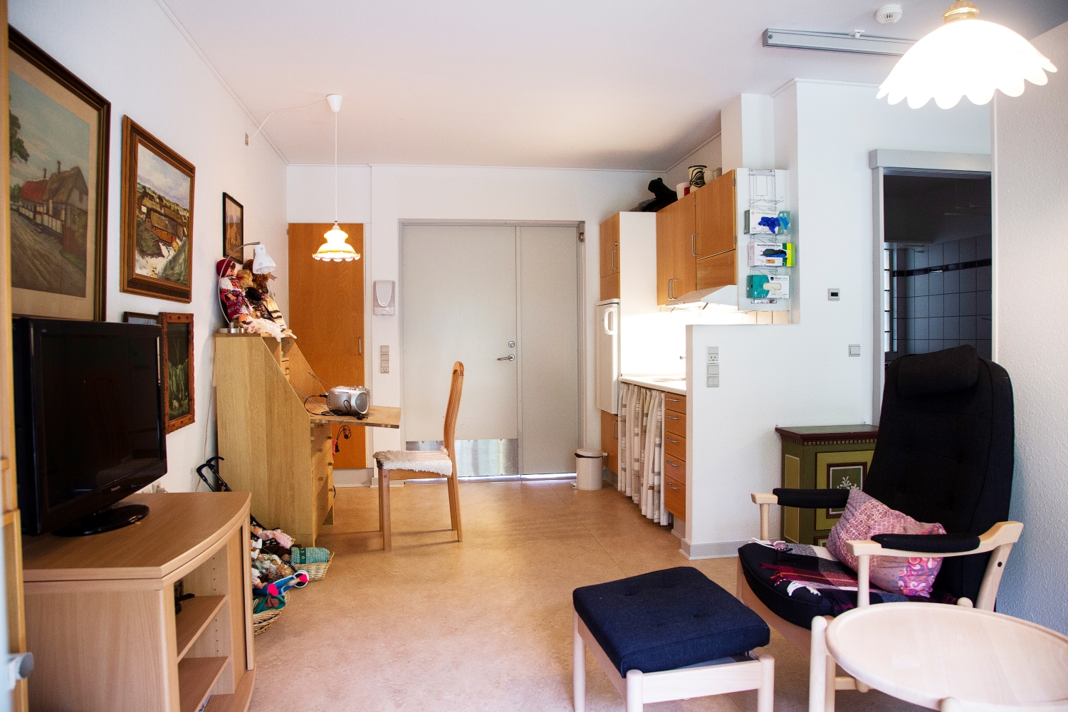 Foran i billedet står der en lænestol med skammel og  kigger over på et fjernsyn som står på en lille reol. På vægen hænger der billeder og i baggrunden er der en lille skrivebord med stol og bagved et lille tekøkken.