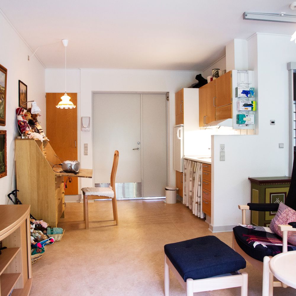 Foran i billedet står der en lænestol med skammel og kigger over på et fjernsyn som står på en lille reol. På vægen hænger der billeder og i baggrunden er der en lille skrivebord med stol og bagved et lille tekøkken.