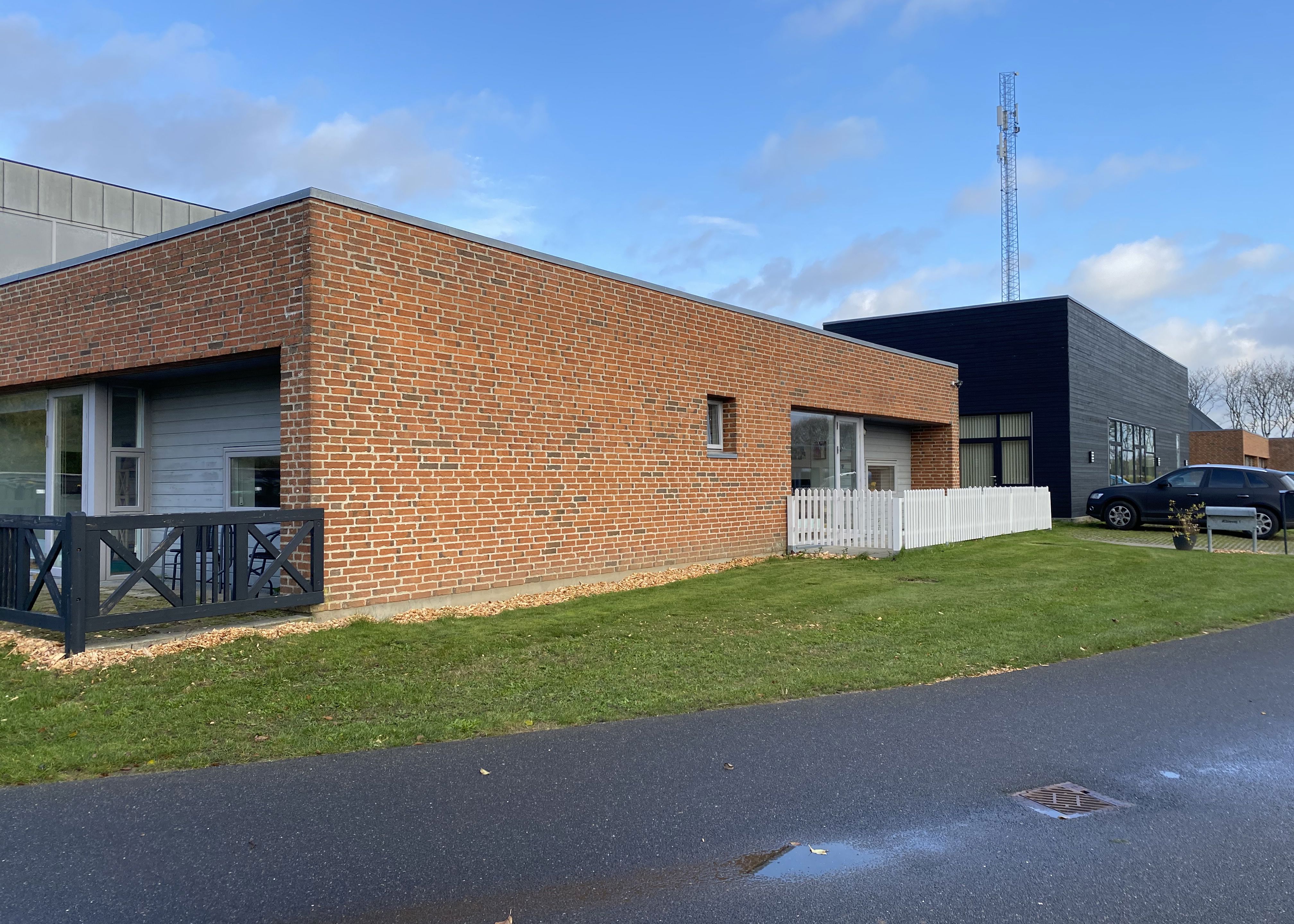 Æblevej 1 udvendigt, der er asfalteret vej udenom. Terrassen bagved Æblevej 1 er indhegnet med et sort lavt stakit. Der er en indhegnet terrasse med et hvidt lavt stakit foran Æblevej 1.