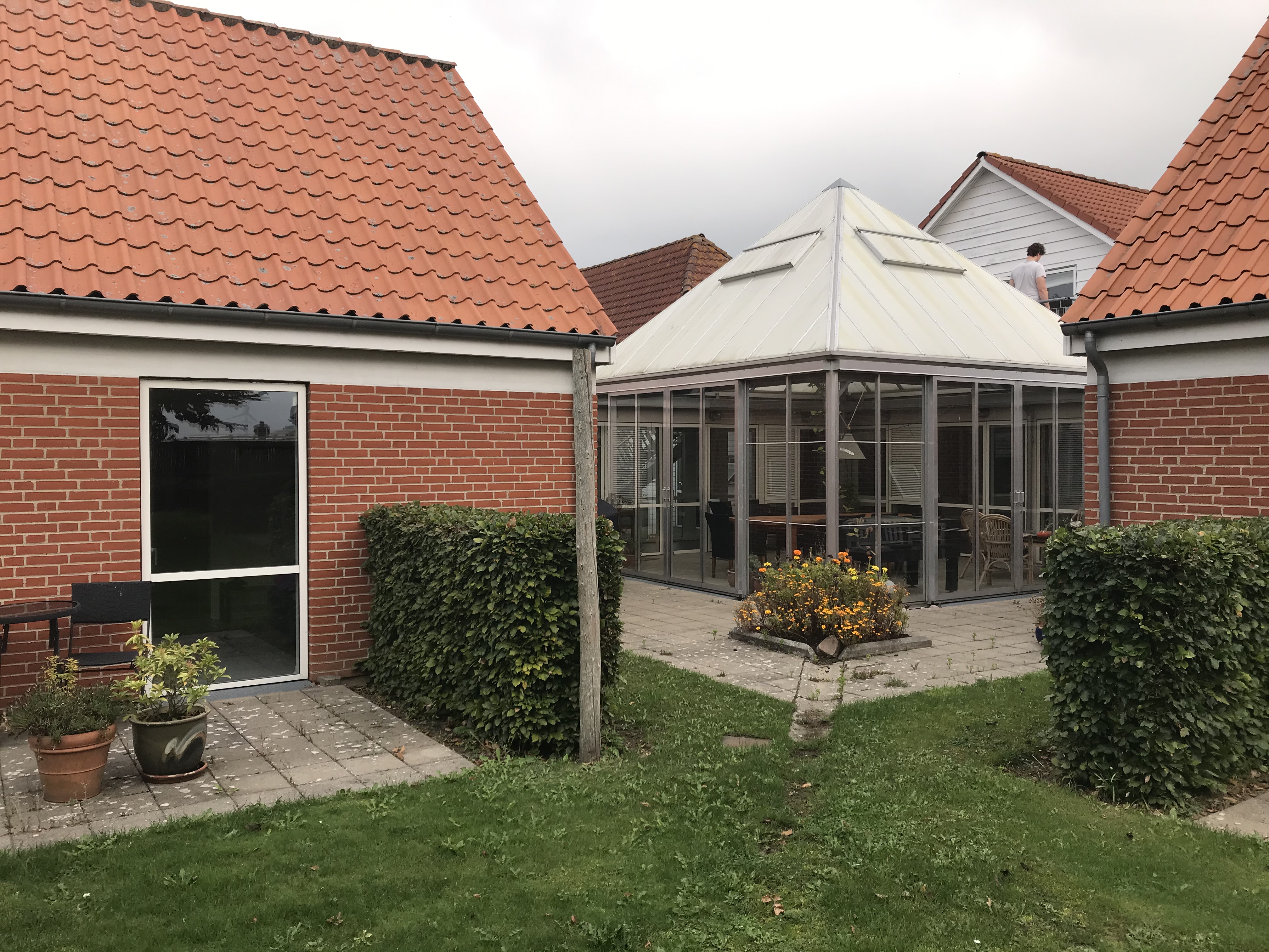 Lærkely set fra haven. En rød bygning med hvide vinduer, en pavilion i  hjørnet af huset, foran er der græs, blomsterbede,  og en terrasse med blomsterkrukker.