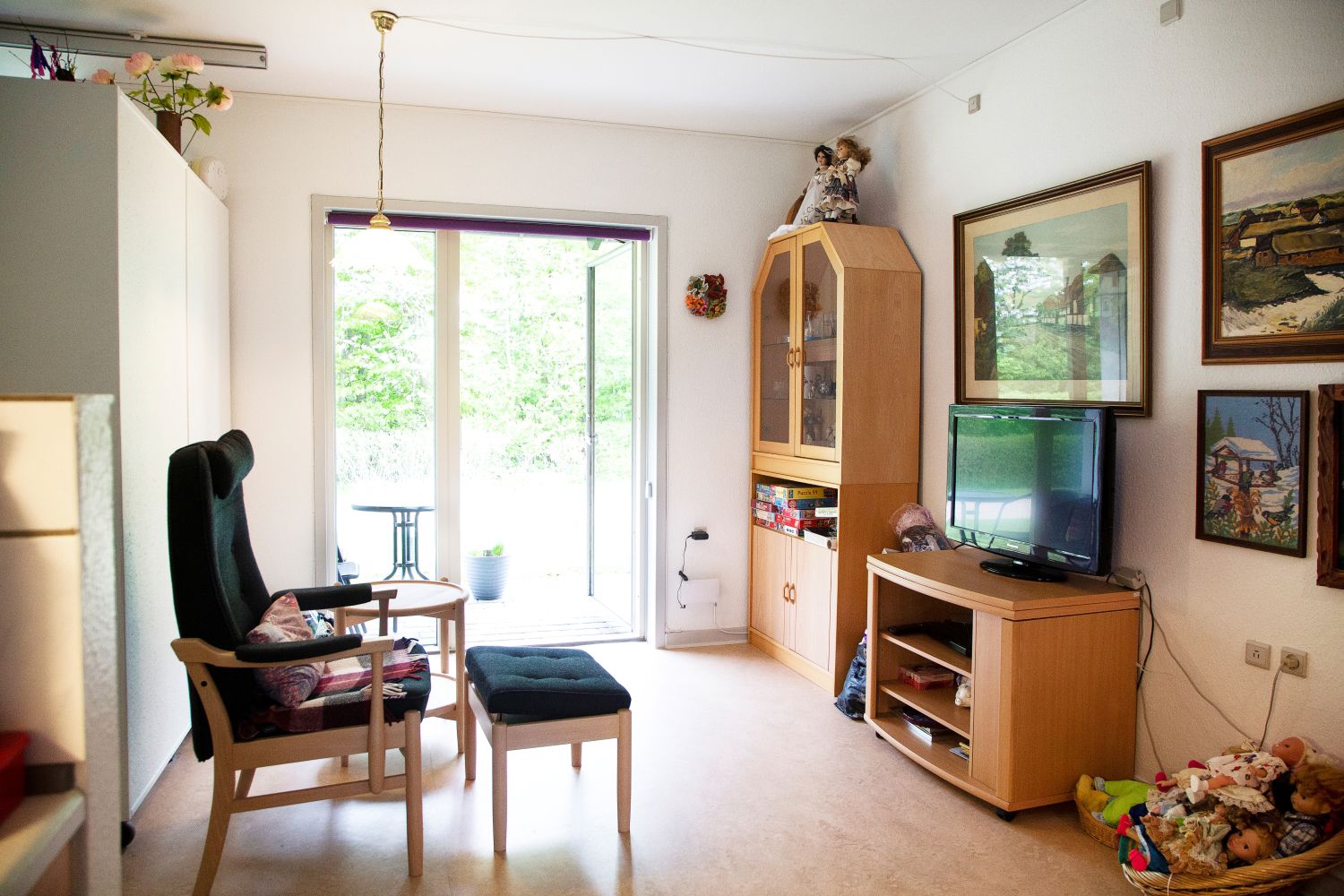 Foran i billedet står der en lænestol med skammel og kigger over på et fjernsyn som står på en lille reol. Og i baggrunden et stort vindue hvor man kigger ud på haven.