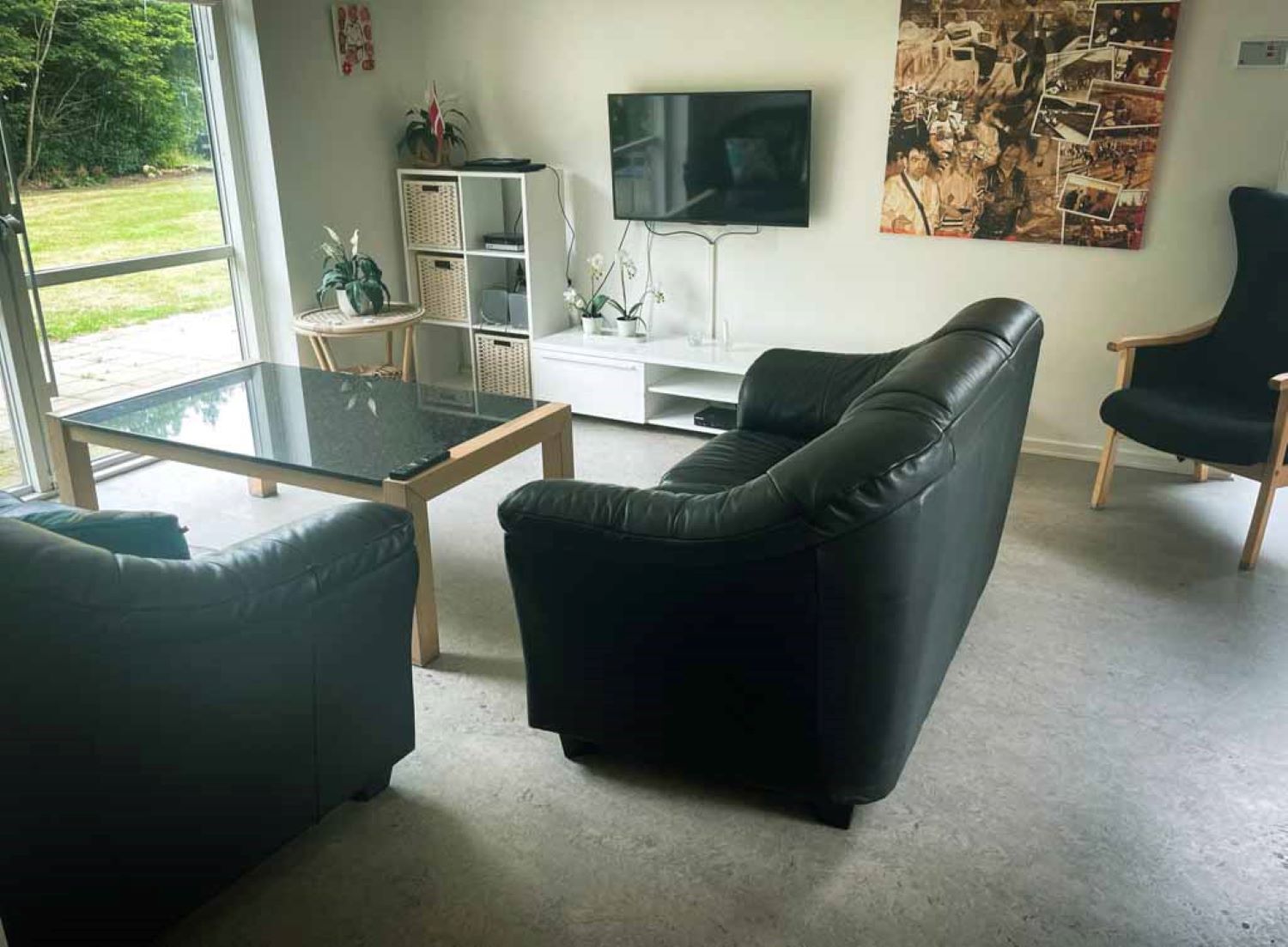 Fællesstue hvor man ser to sofaer i sort læder et sofabord, på væggen hænger der et fjernsyn og under det en lille hvidreol.