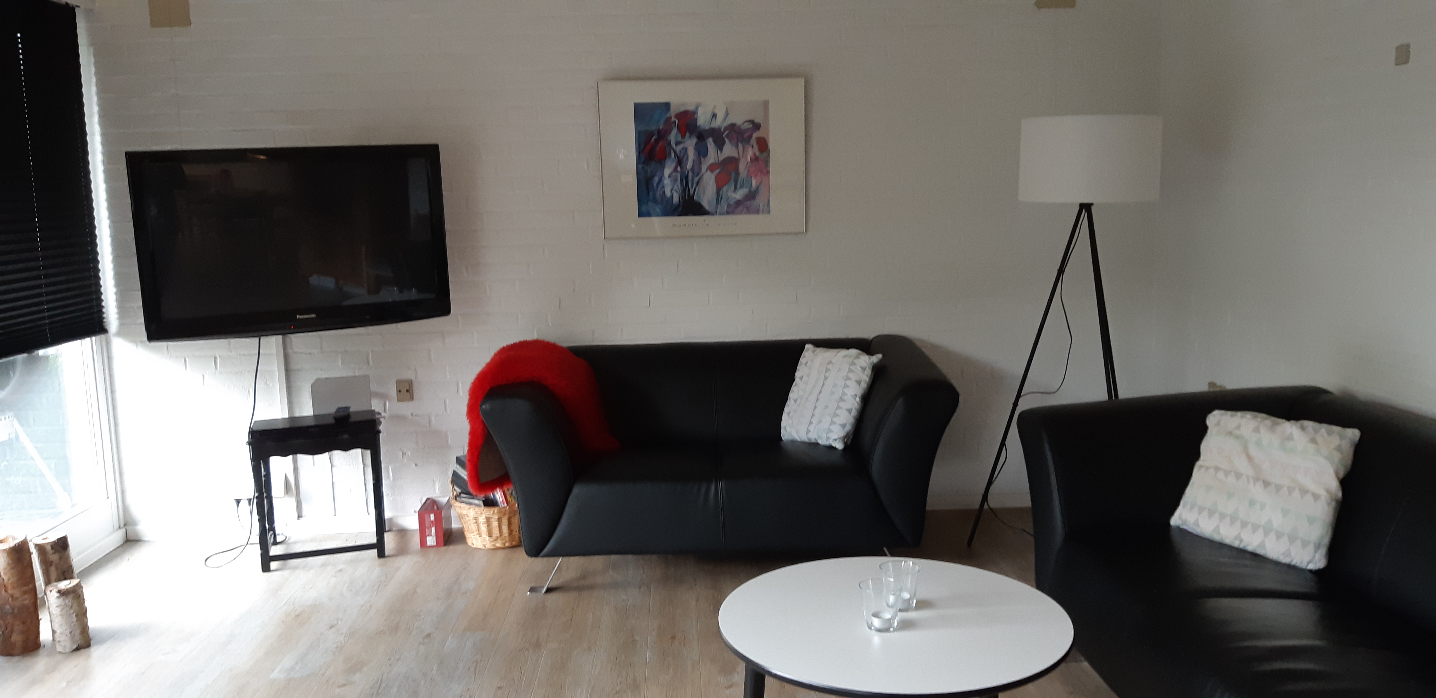 Opholdsstue med to lædersofaer, et rundt bord og et tv. I hjærnet er der er høj standerlampe med hvid lampeskærm. 