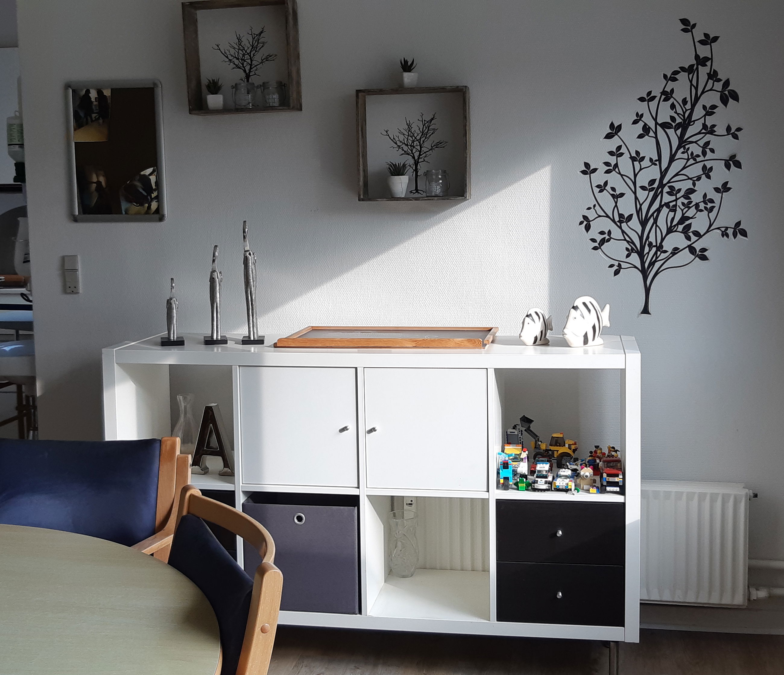 Fællesrummet hvor der er et køkken i baggrunden og et rundt spisebord med stole og en reol ved siden af.