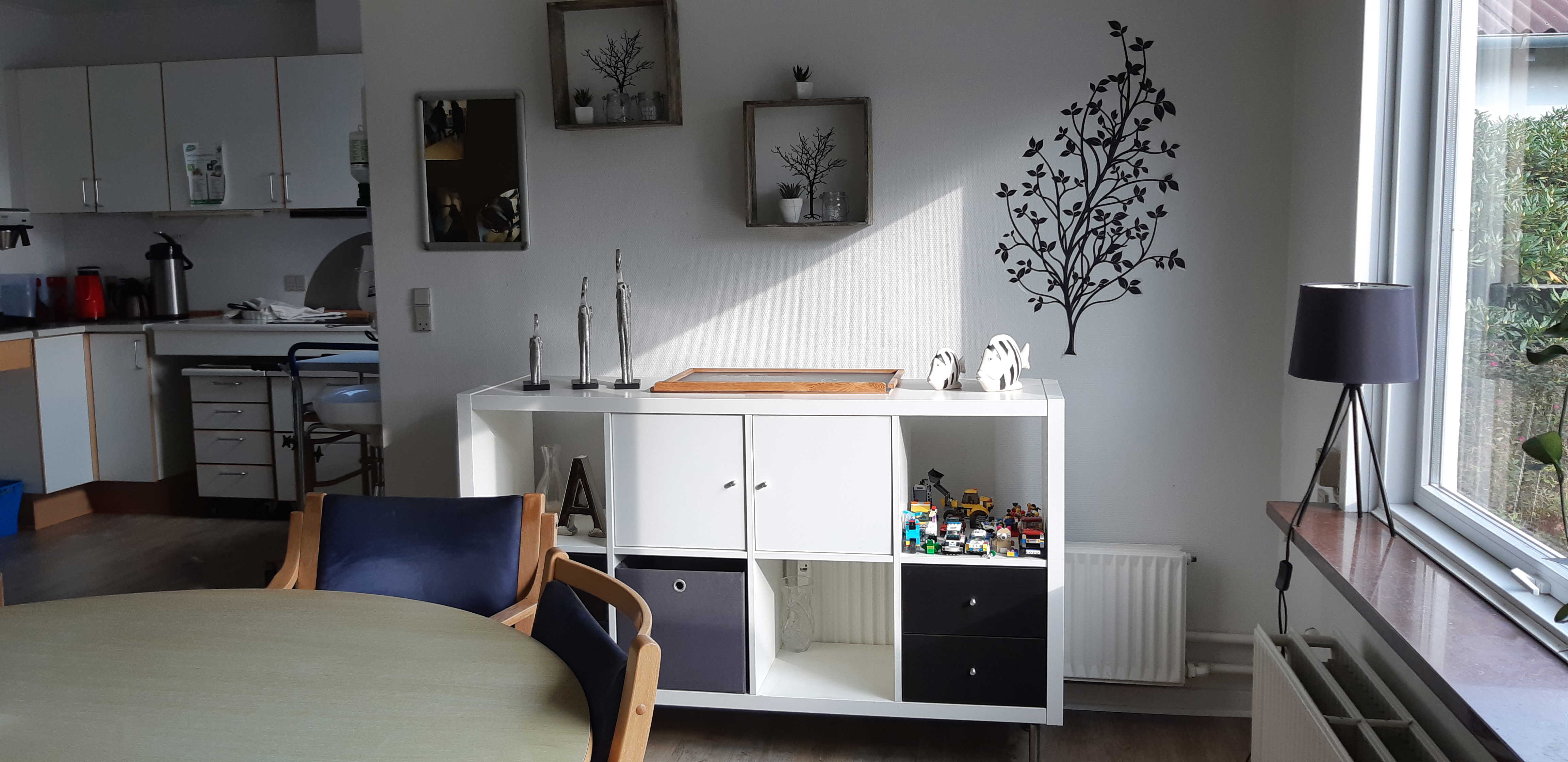 Fællesrummet hvor der er et køkken i baggrunden og et rundt spisebord med stole og en reol ved siden af.