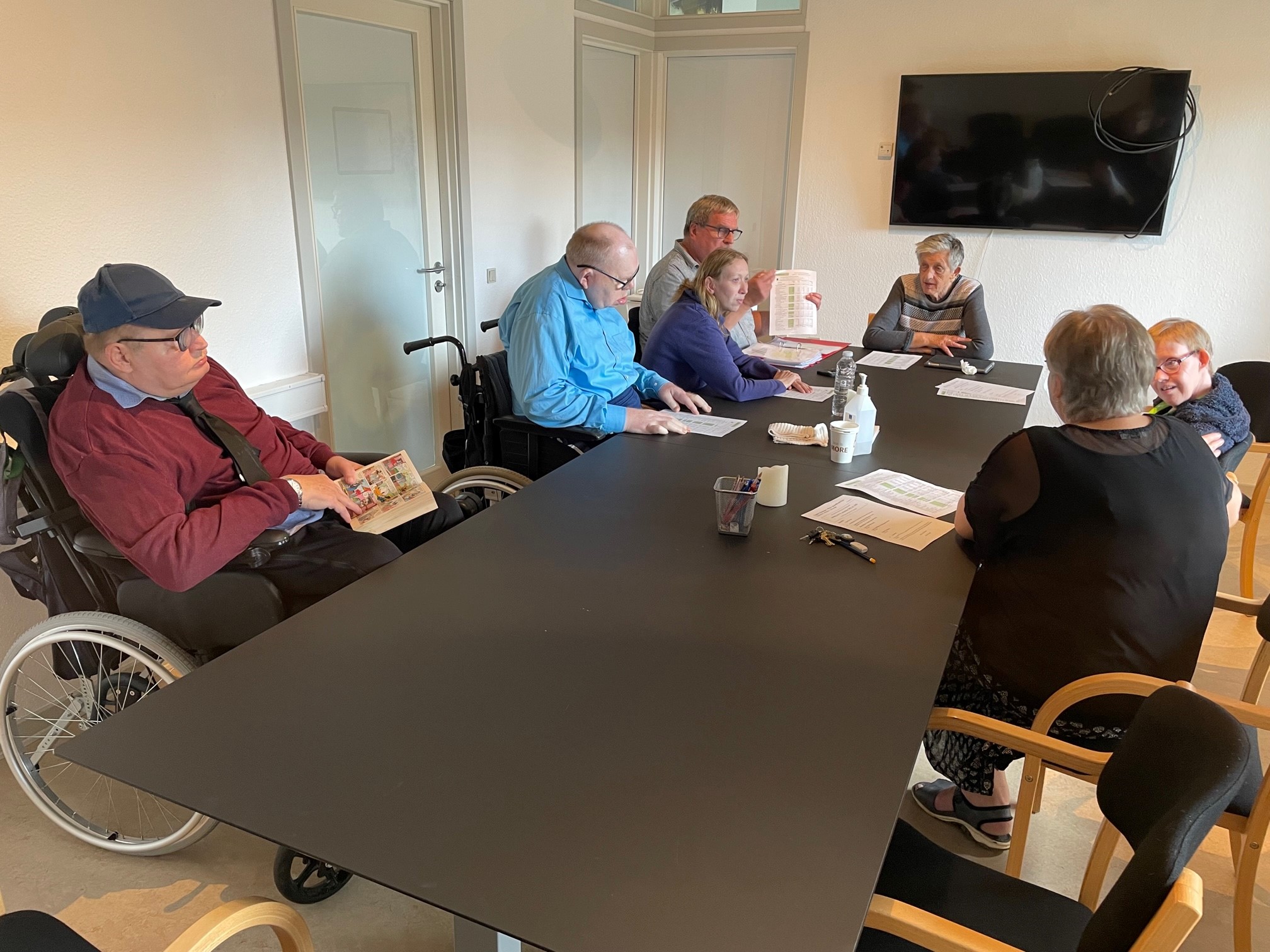 Et langt bord hvor der sidder syv personer omkring som deltager i afdelingsmøde.