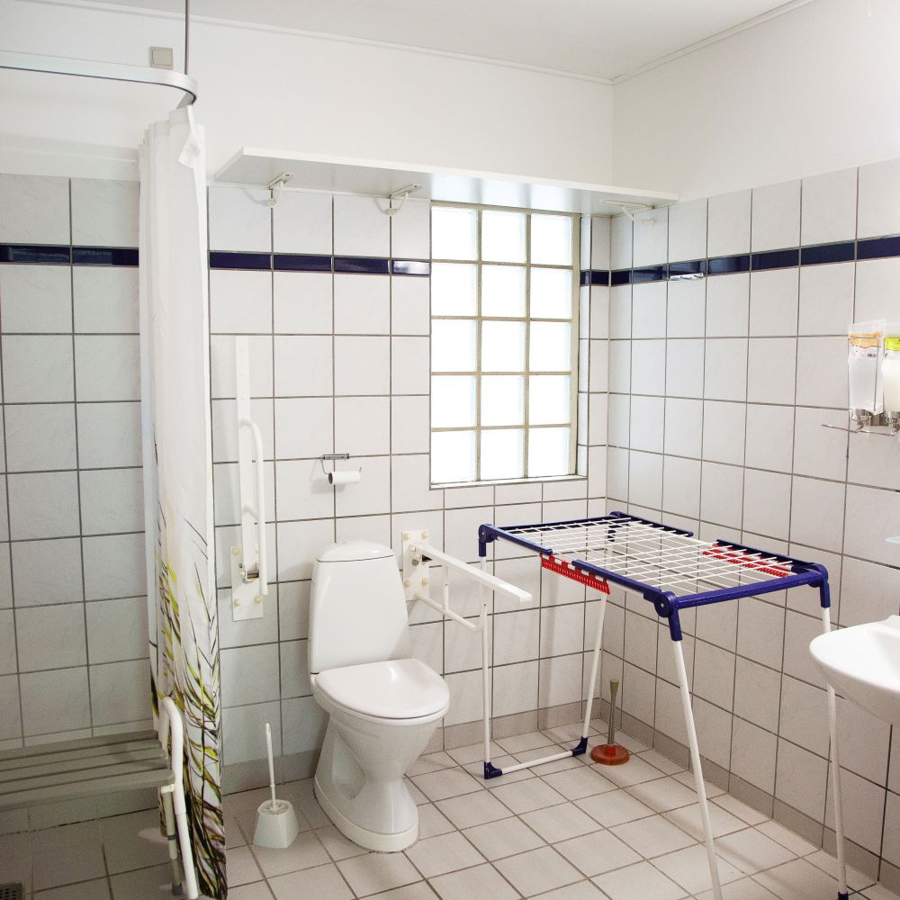 Badeværelse med toilet og vask og et brusehjørne med forhæng og i hjørnet står der et tøjstativ.