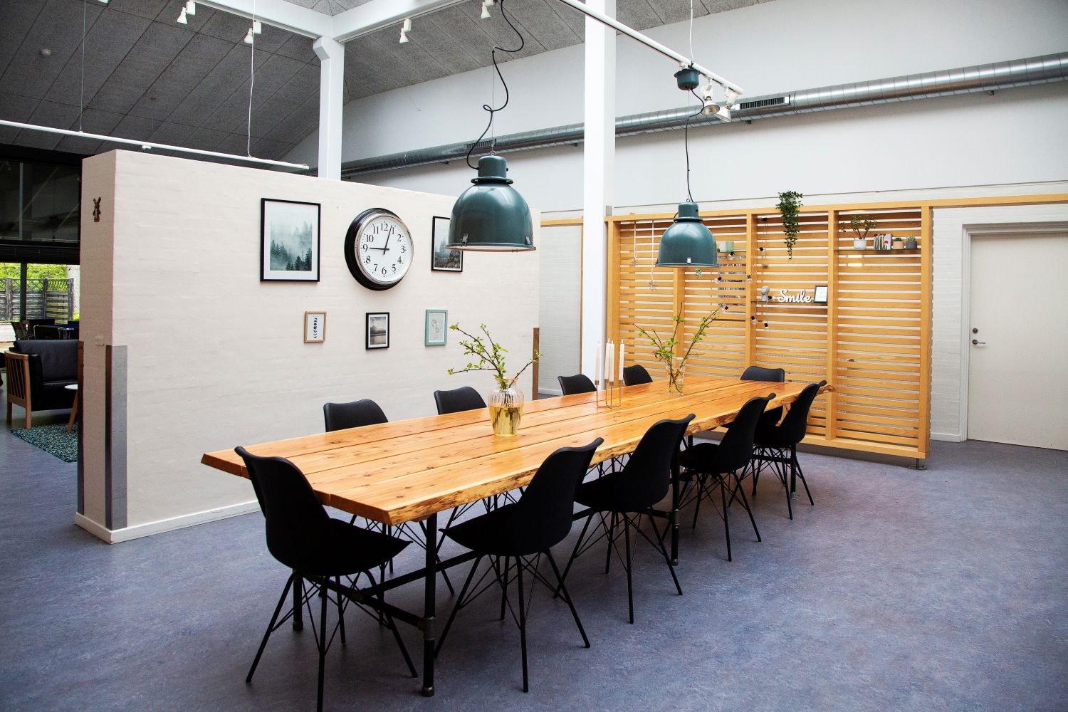 Træspisebord af lakeret planker gulvet er blåt stolene er sorte bagved en halvmur der er hvid for enden af bordet er en skillevæg i træ med forskelligt pynt på