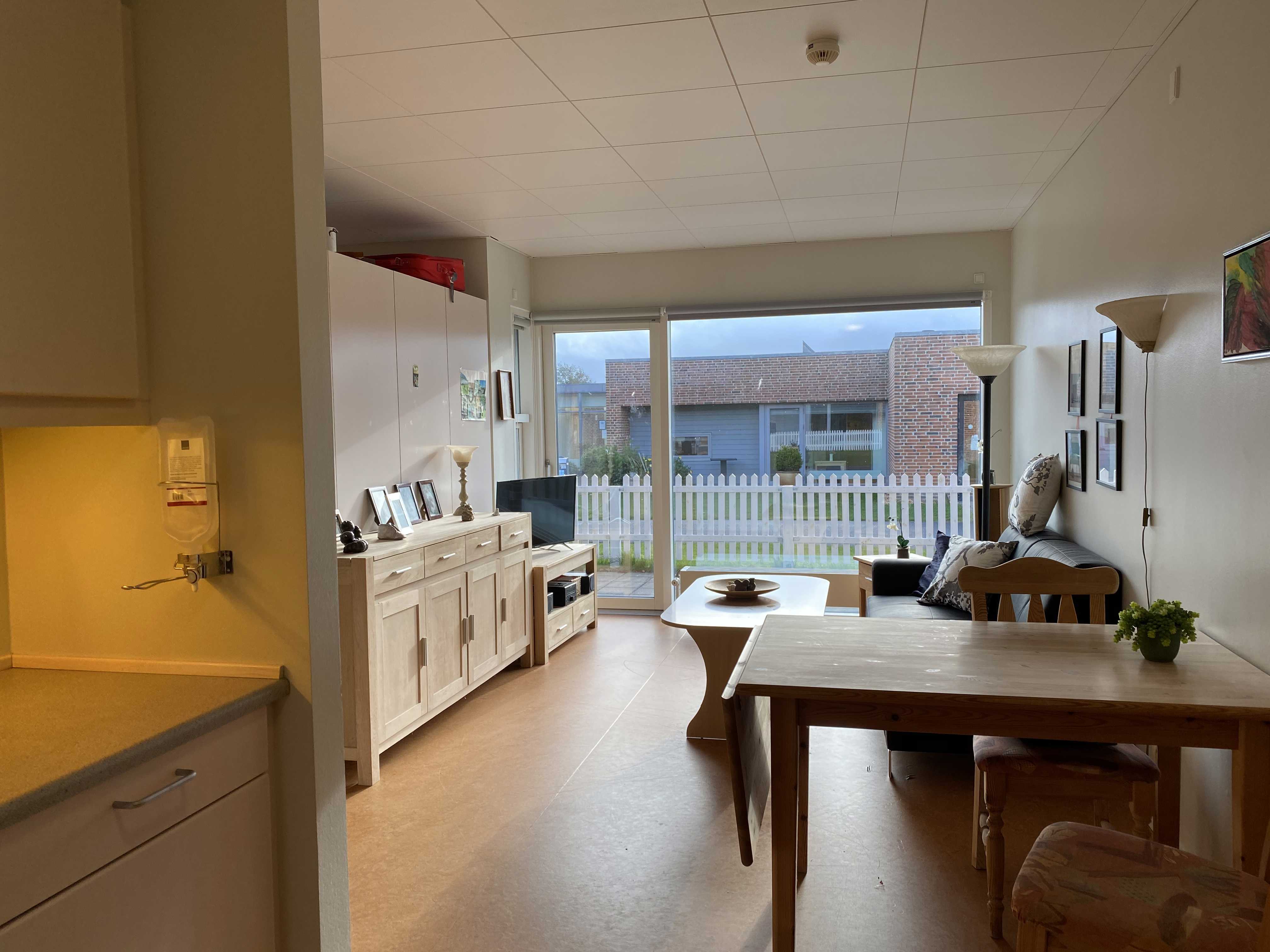 Inde i en borgers lejlighed er der en sofa og et sofabord. I lejligheden der en skænk og et tv bord. I lejligheden er der også et spisebord og et lille thekøkken.