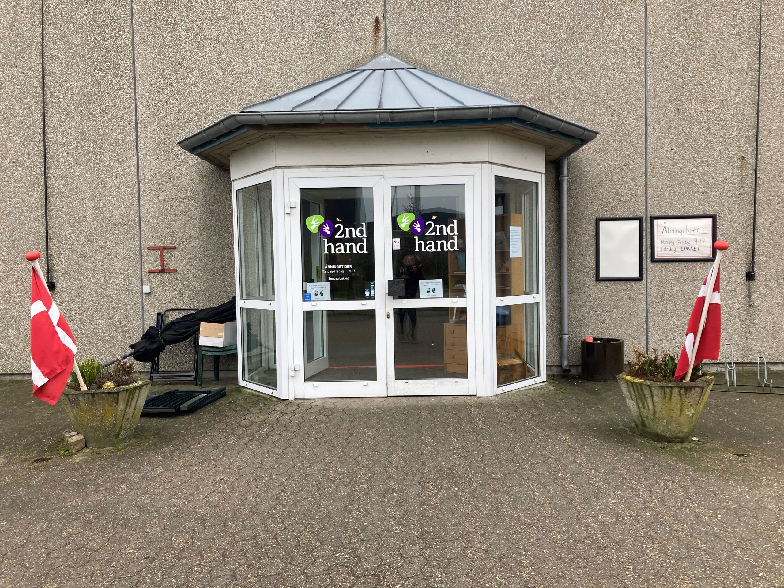 Indgangspartiet til 2Nd Hand butikken, det er et glasdørsparti hvor der står 2Nd Hand med grøn og lilla, foran er der to beton kummer med lyng i og flag