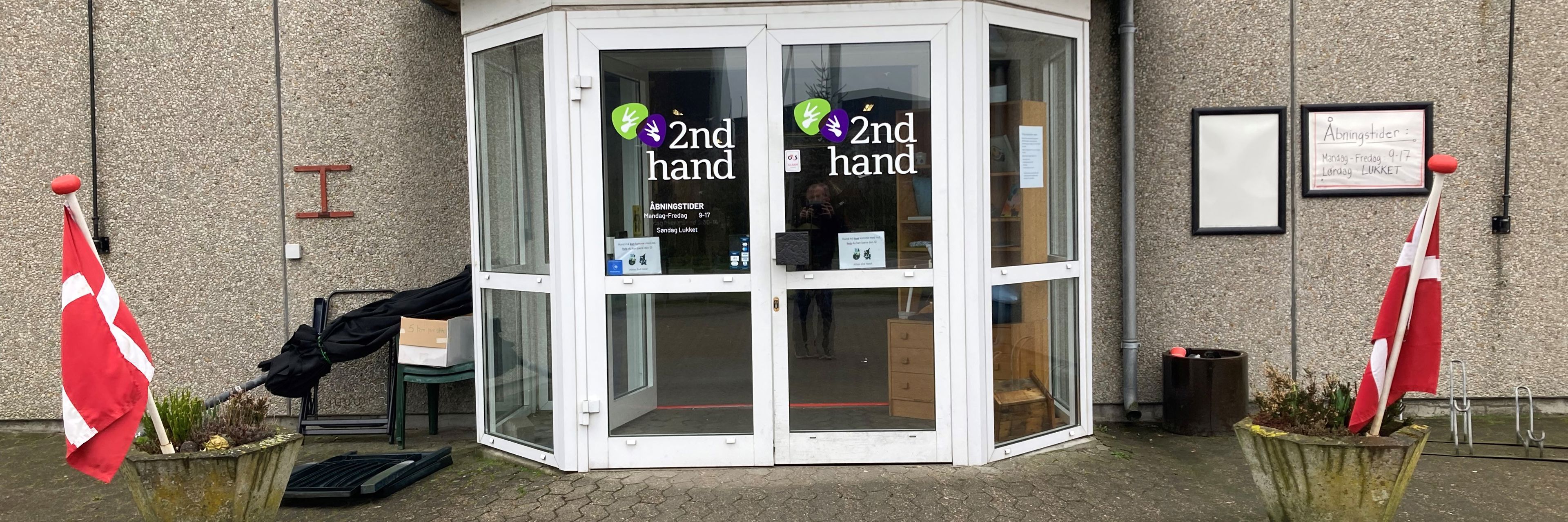 Indgangspartiet til 2Nd Hand butikken, det er et glasdørsparti hvor der står 2Nd Hand med grøn og lilla, foran er der to beton kummer med lyng i og flag