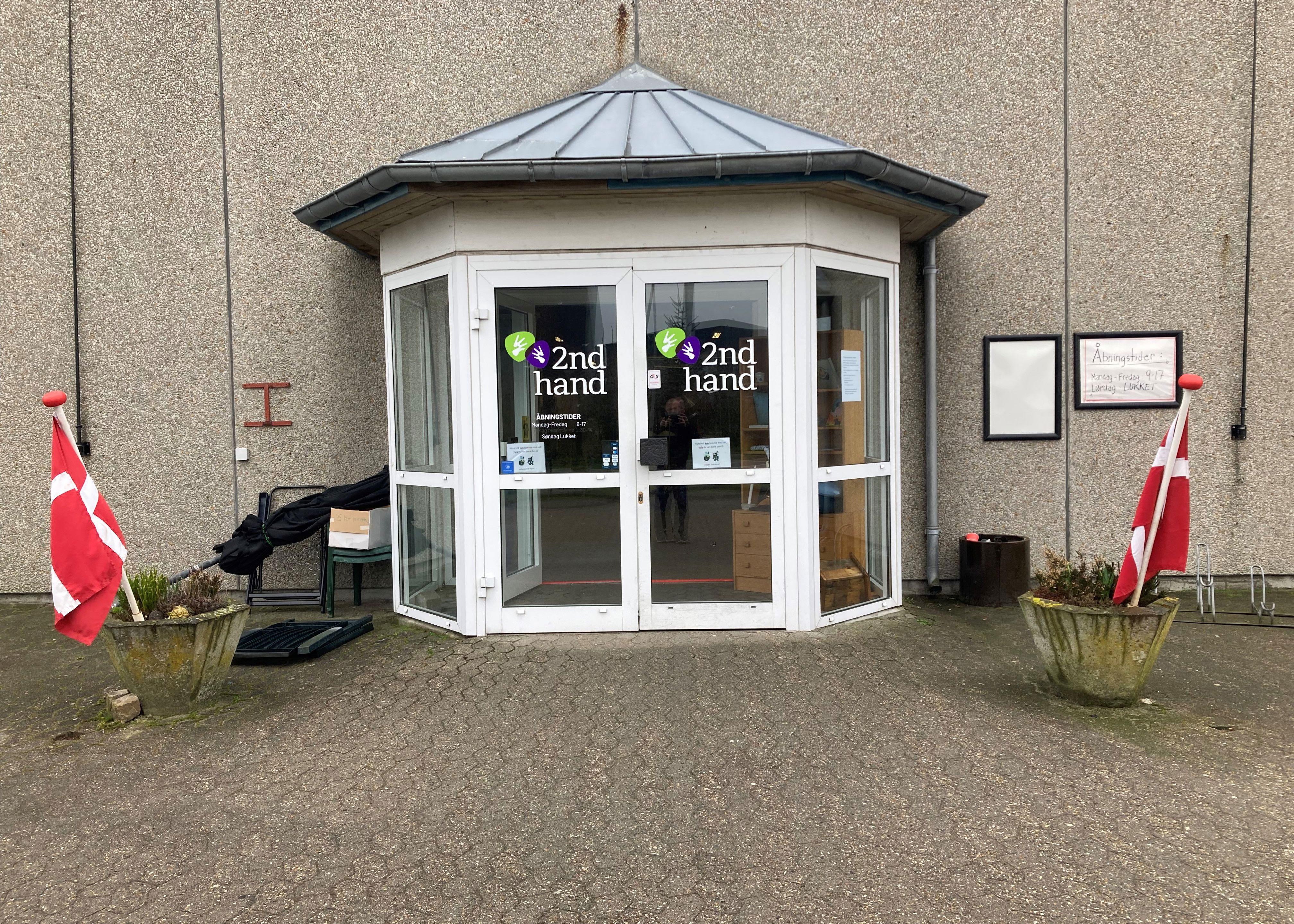Indgangspartiet til 2Nd Hand butikken, det er et glasdørsparti hvor der står 2Nd Hand med grøn og lilla, foran er der to beton kummer med lyng i og flag