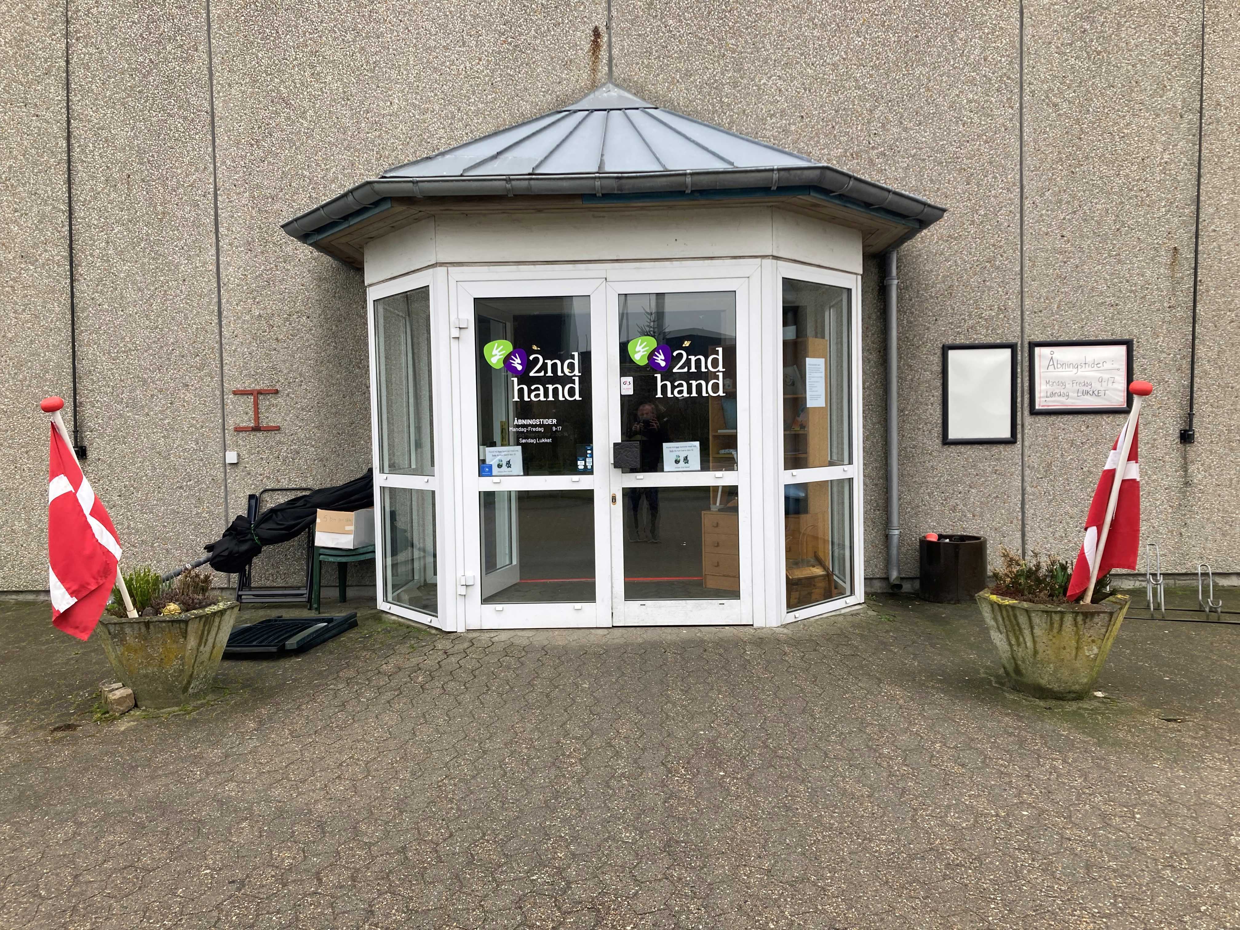 Indgangspartiet til 2Nd Hand butikken, det er et glasdørsparti hvor der står 2Nd Hand med grøn og lilla, foran er der to beton kummer med lyng i og flag