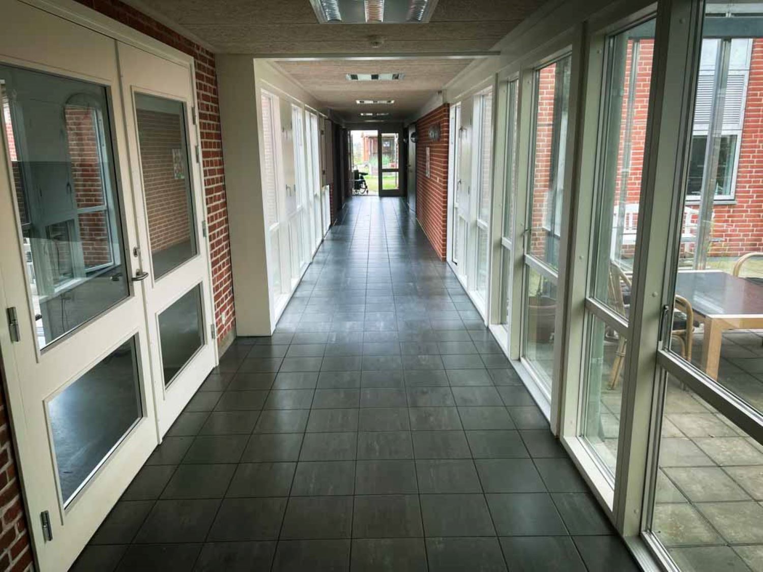 En lang gang med hvide vinduer og døre og gulvet er af mørke fliser og for enden af gangen er der en dør ud til haven.