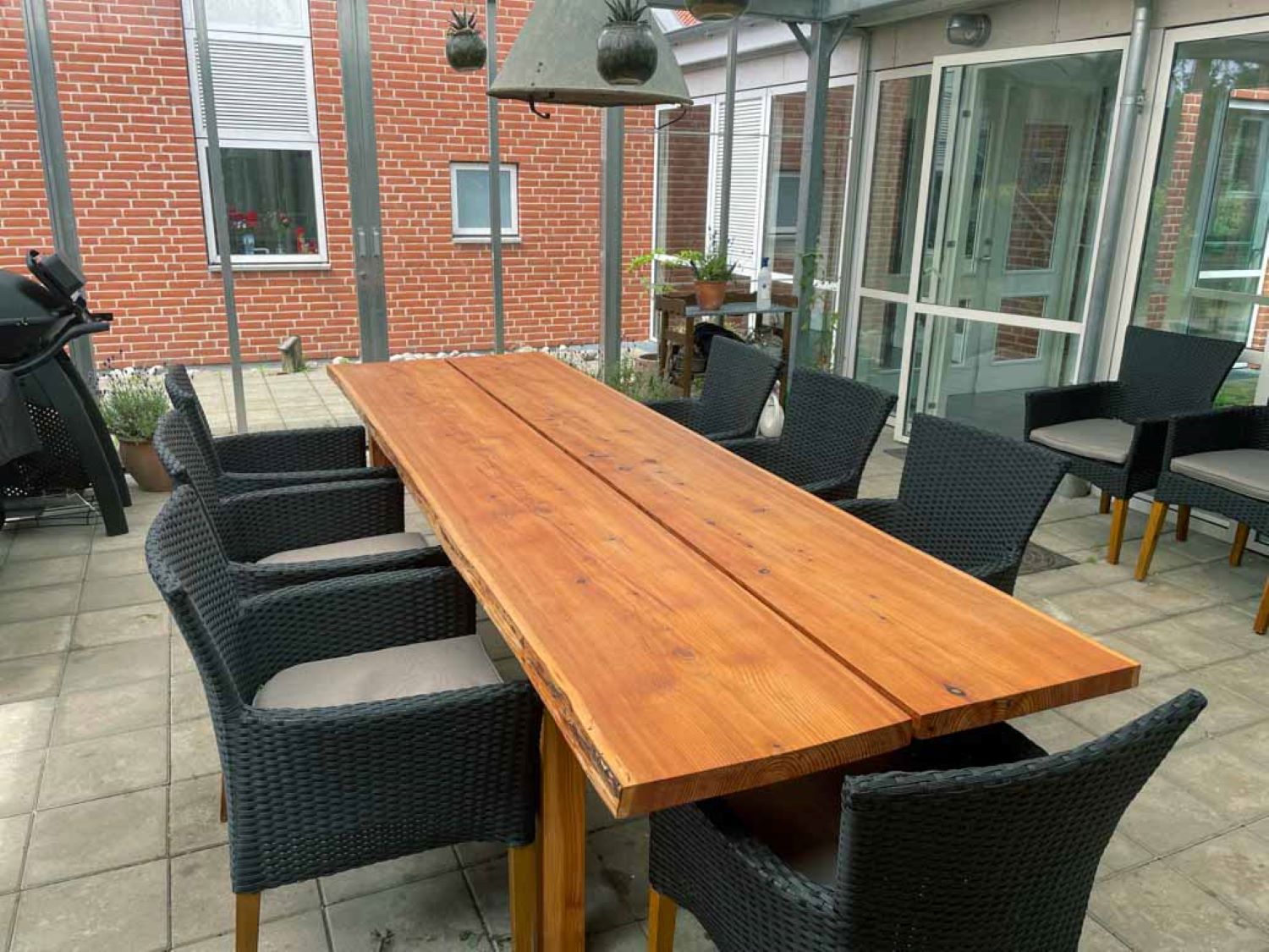 I pallionen står en langt plankebord med syv sorte havestole omkring, i den ene hjørne kan man se en gasgril og der står to stole i baggrunden.