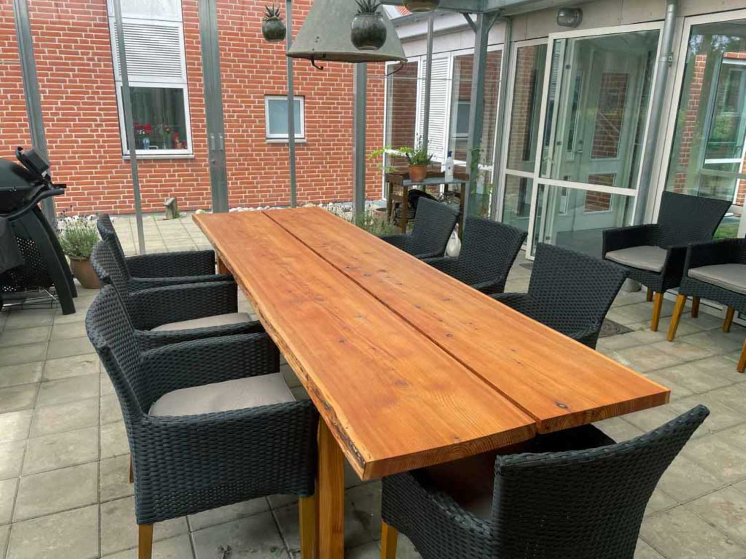 I pallionen står en langt plankebord med syv sorte havestole omkring, i den ene hjørne kan man se en gasgril og der står to stole i baggrunden.