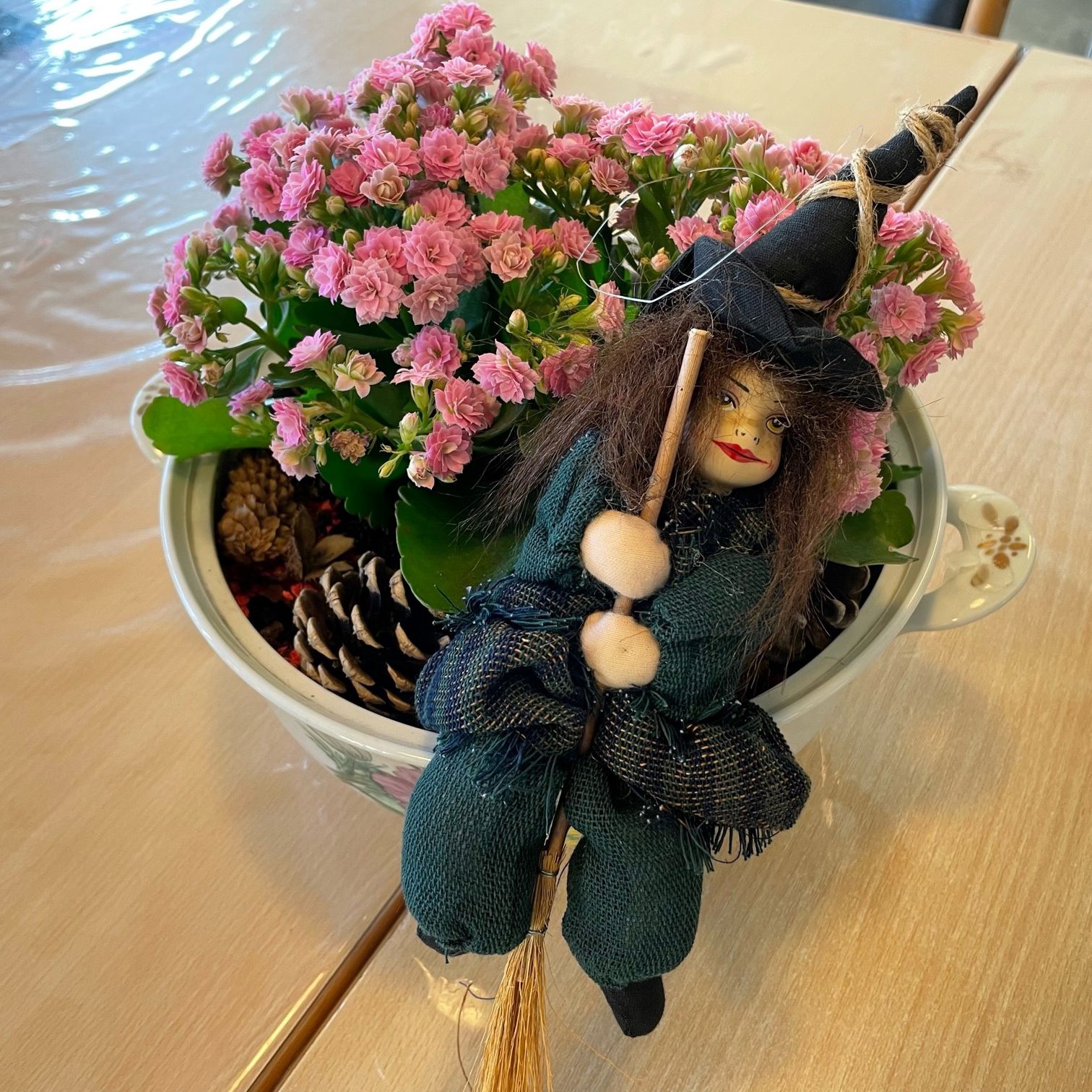 En blomster dekoration med lyserøde blomster i, grankogler og en pynteheks den står på et træbord med en gennemsigtig voksdug på