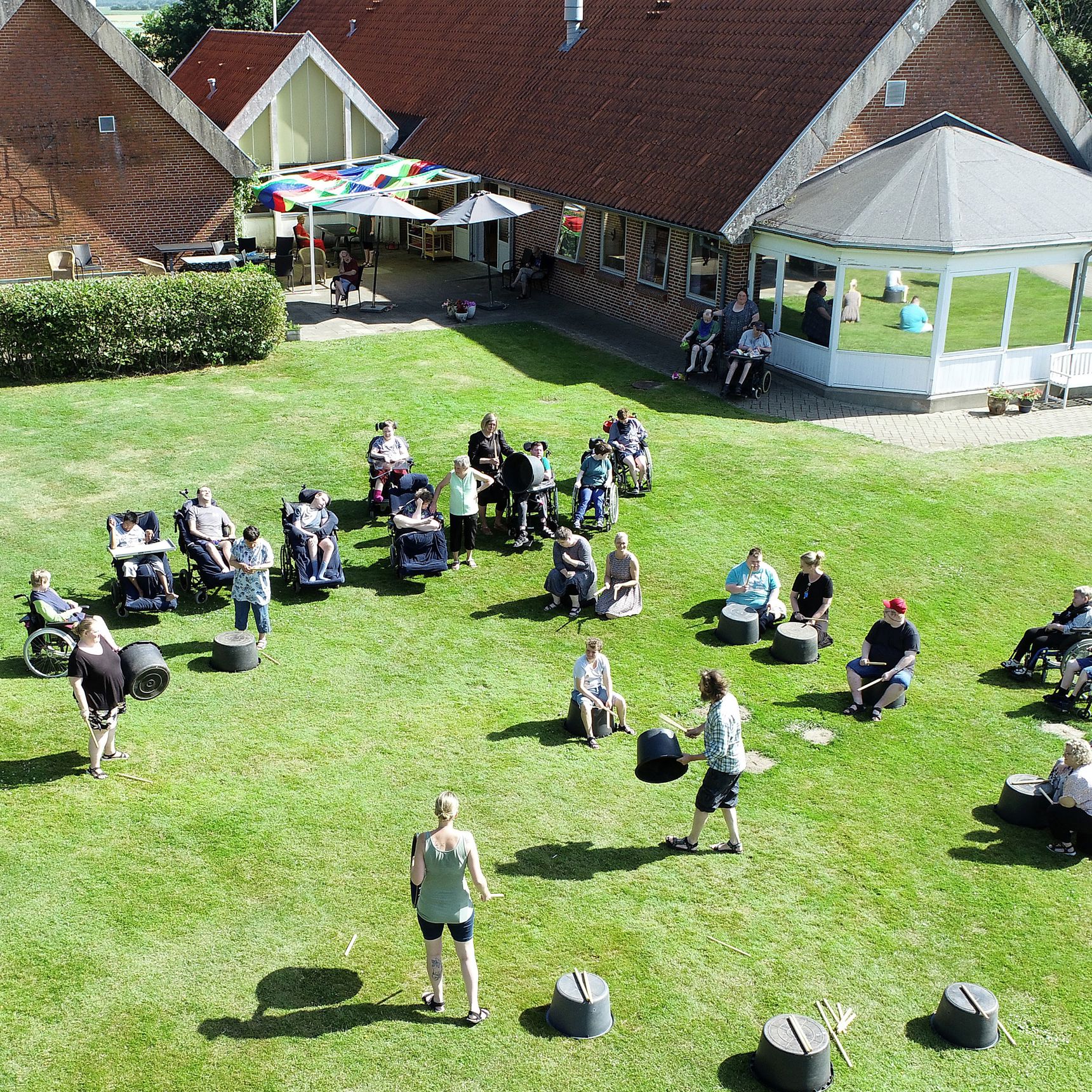 Foto taget oppefra af borgere der står spredt på en græsplæne med nogle baljer, nogle står på baljerne med trommestikker, bagved ses bygningen med rode mursten og et faverigt soldækken over terrassen.