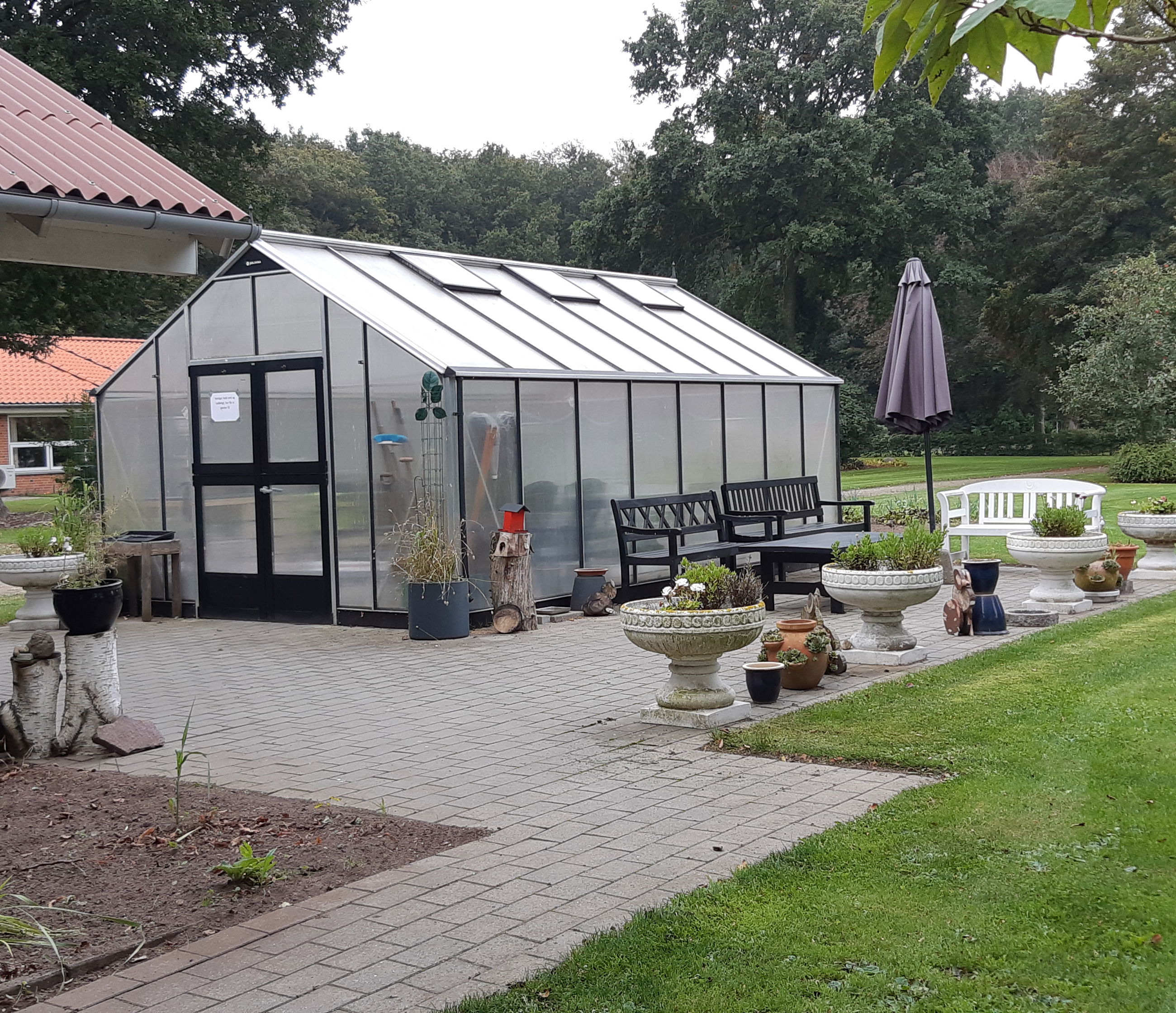 Der er et stort drivhus som ligger på en flisebelagt grund og rundt om er der en stor græsplæne, der er bænke hvor man kan sidde.