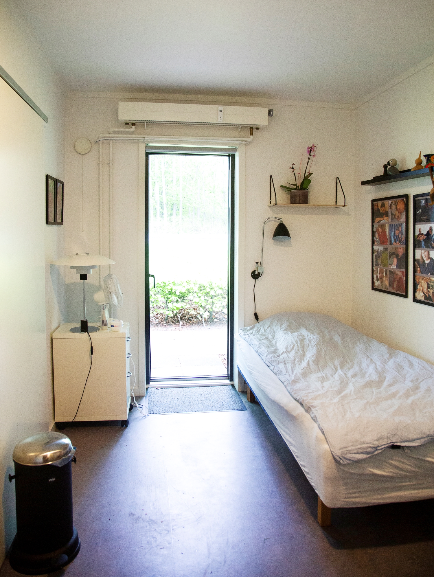 soveværelse i lejlighed hvide vægge en seng med hvidt sengetøj en havedør for enden af rummet og overfor en kommode med en lampe på