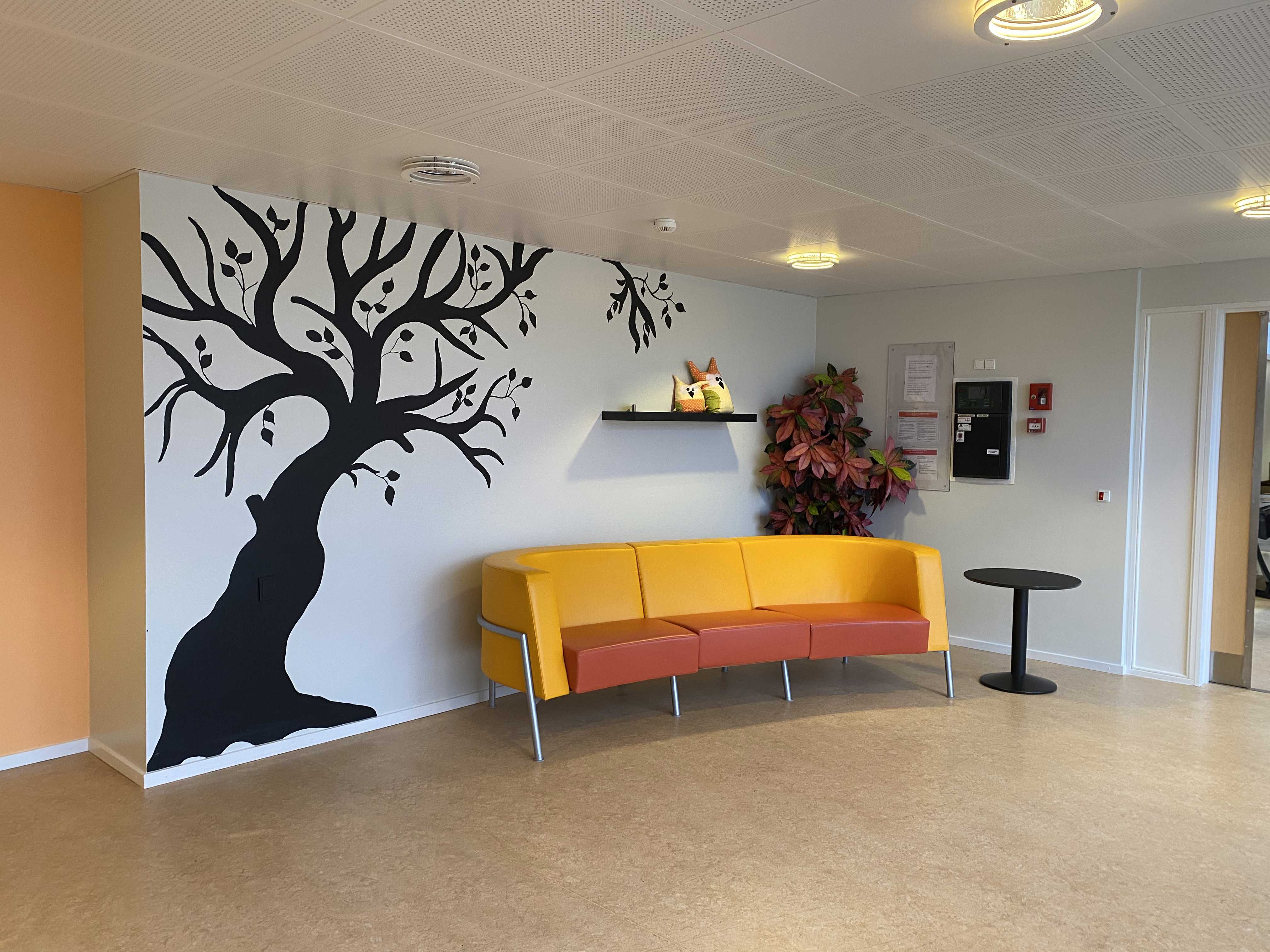 Indgangen på Æblevej 1. På væggen bagerst i billedet er der malet et stort sort træ på væggen, lige foran væggen står der en gul og orange læder sofa, og til højre for den et lille sort cafebord. Gulvet er lyst laminat.