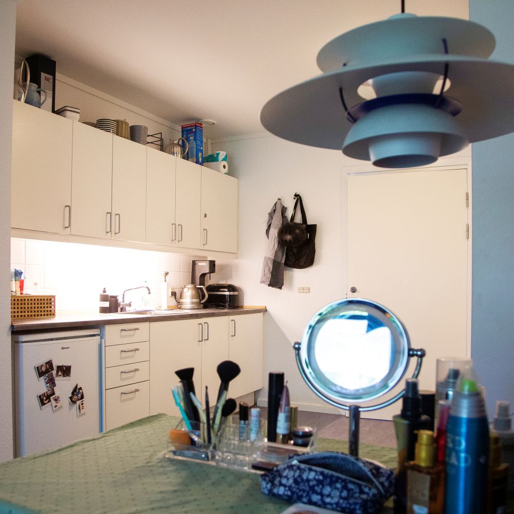 Køkken i lejlighed hvidt med billeder på køleskabet foran står et bord med kosmetik et spejl og hårprodukter