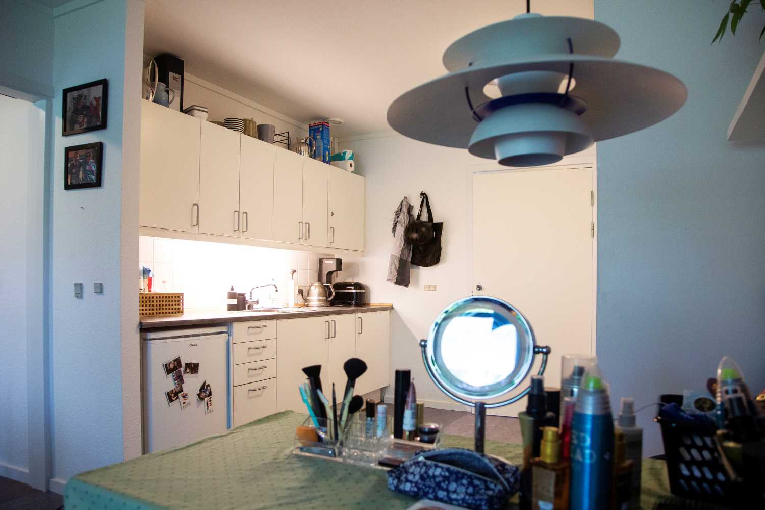 Køkken i lejlighed hvidt med billeder på køleskabet foran står et bord med kosmetik et spejl og hårprodukter